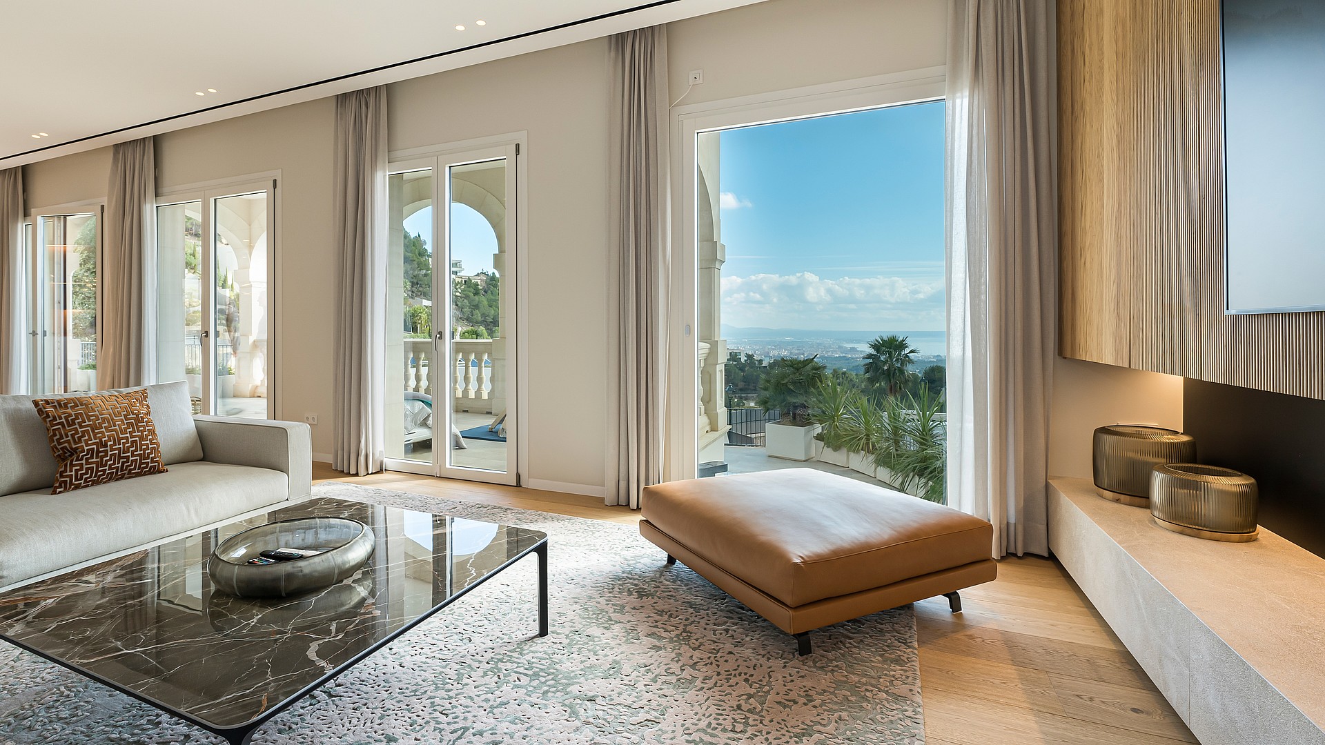 A modern living room with a neutral sofa, glass coffee table, and leather ottoman facing floor-to-ceiling balcony doors.