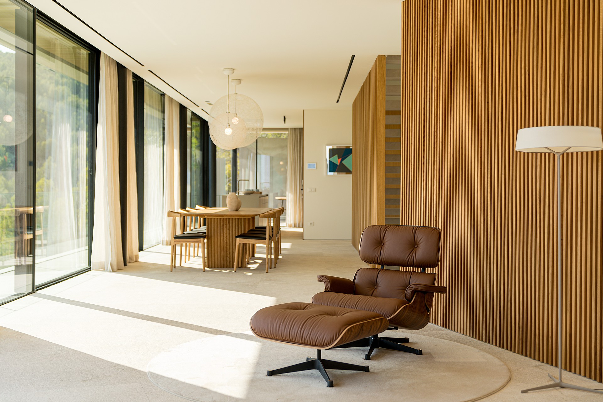 A bright living space featuring a brown leather lounge chair, wooden dining table, and large glass walls looking outside.