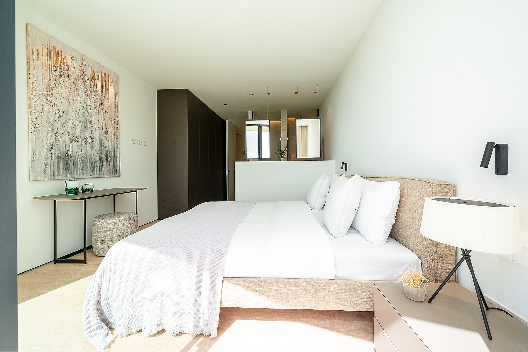 A modern master bedroom with a king-sized bed, white linens, a textured headboard, a console table with abstract art, and an open en-suite bathroom area.