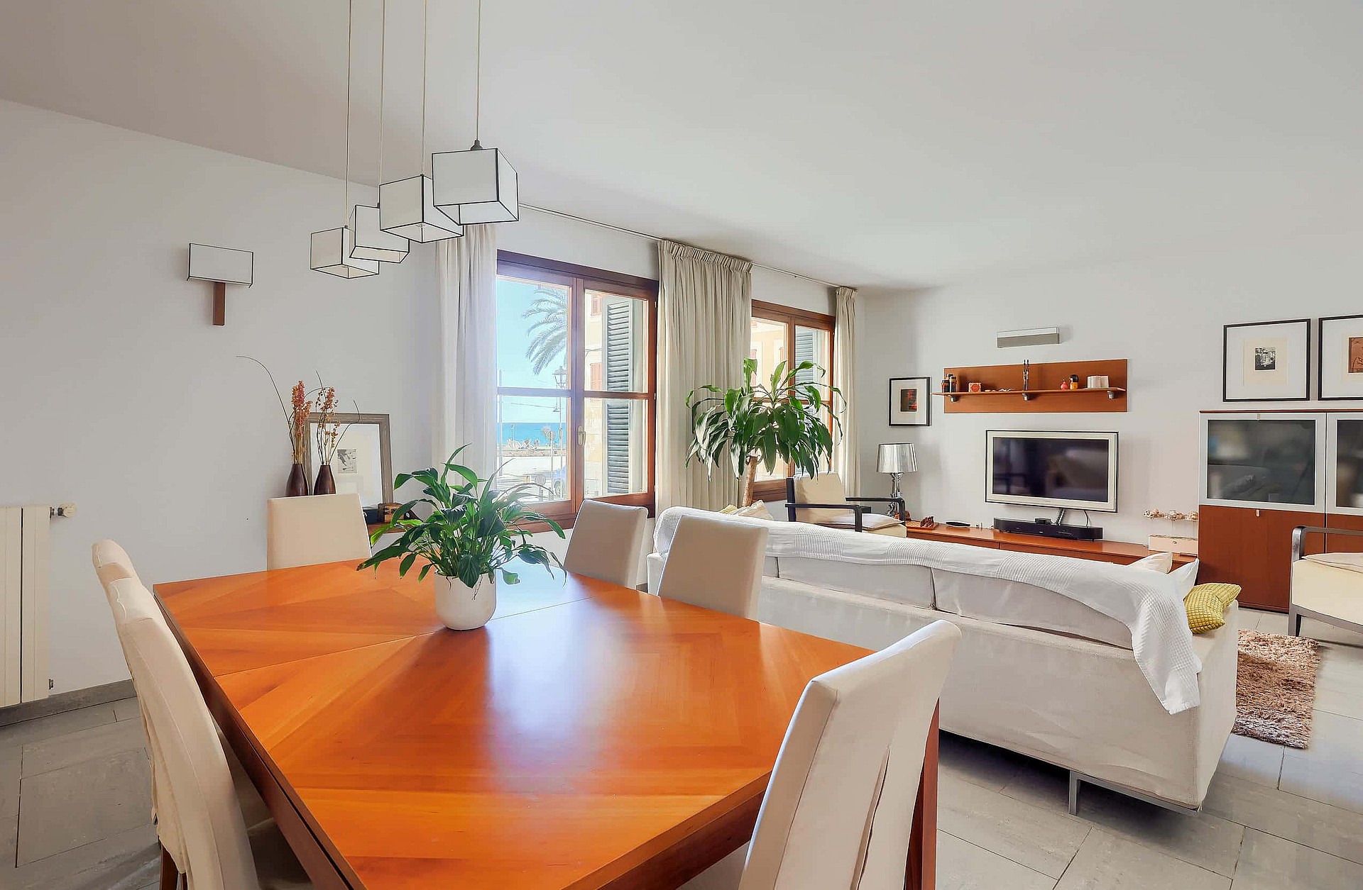 A bright living and dining room with wooden furniture, white sofas, and windows overlooking the sea.