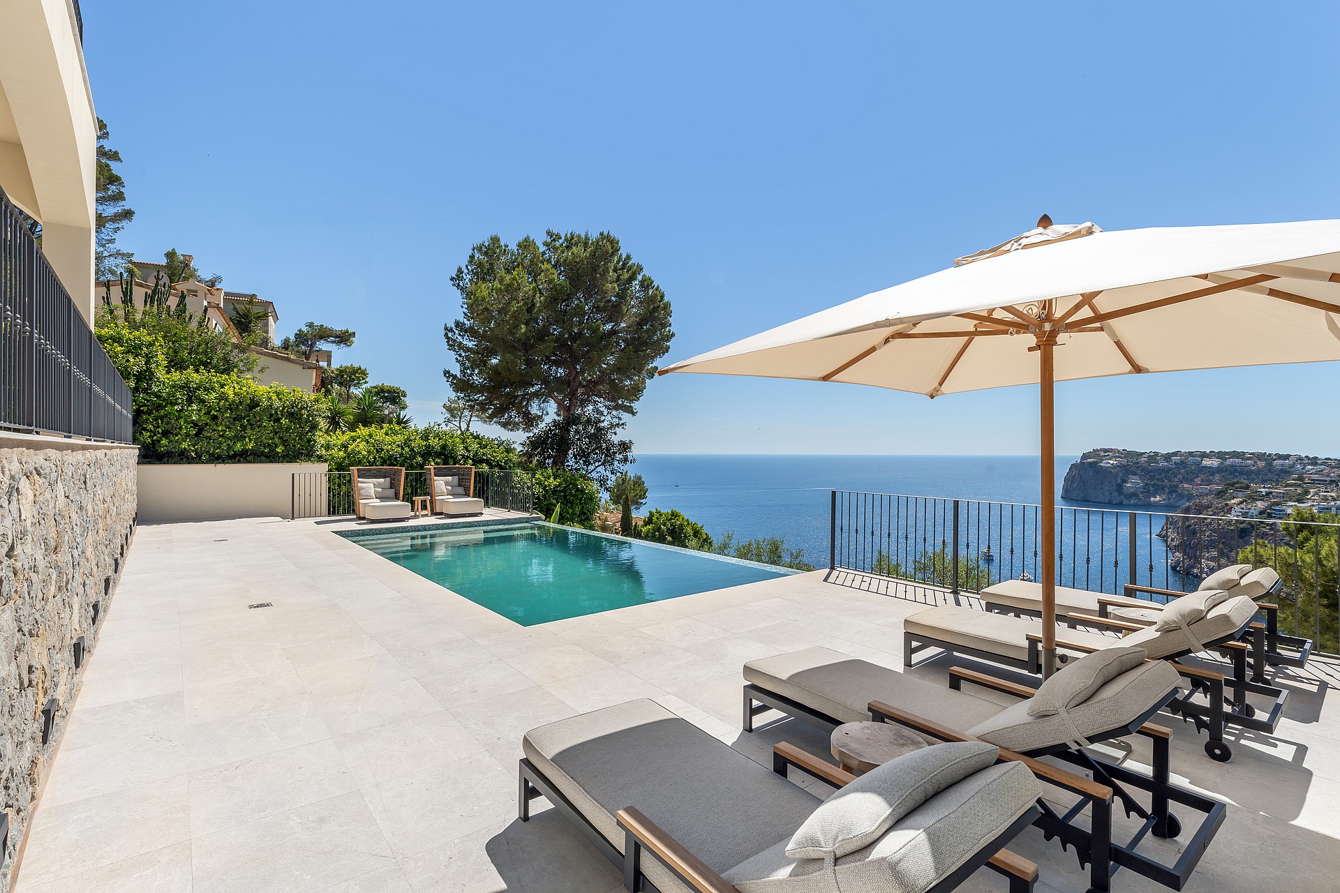 Infinity pool on a light stone terrace with sun loungers overlooking the blue sea under a clear sky.