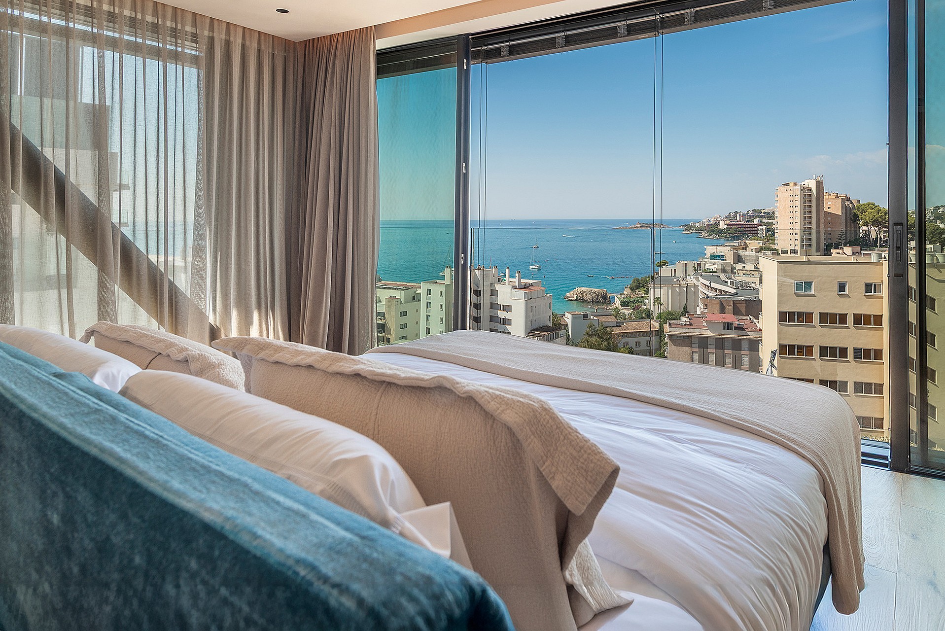 A view from inside a bedroom looking through floor-to-ceiling glass sliding doors at a Mediterranean coastline and sea.