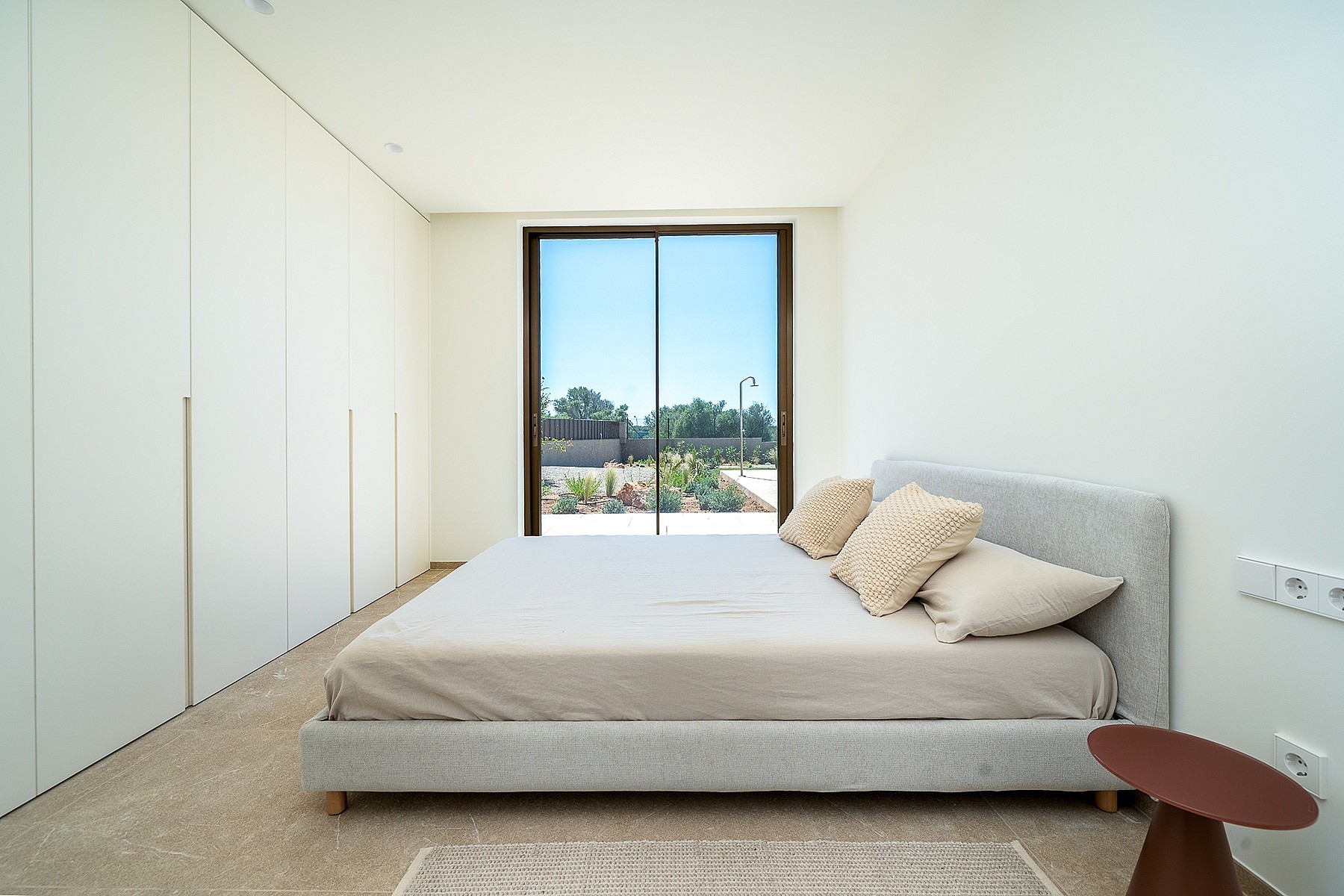A minimalist bedroom featuring a grey upholstered bed, floor-to-ceiling white wardrobe, and a sliding glass door to a garden.