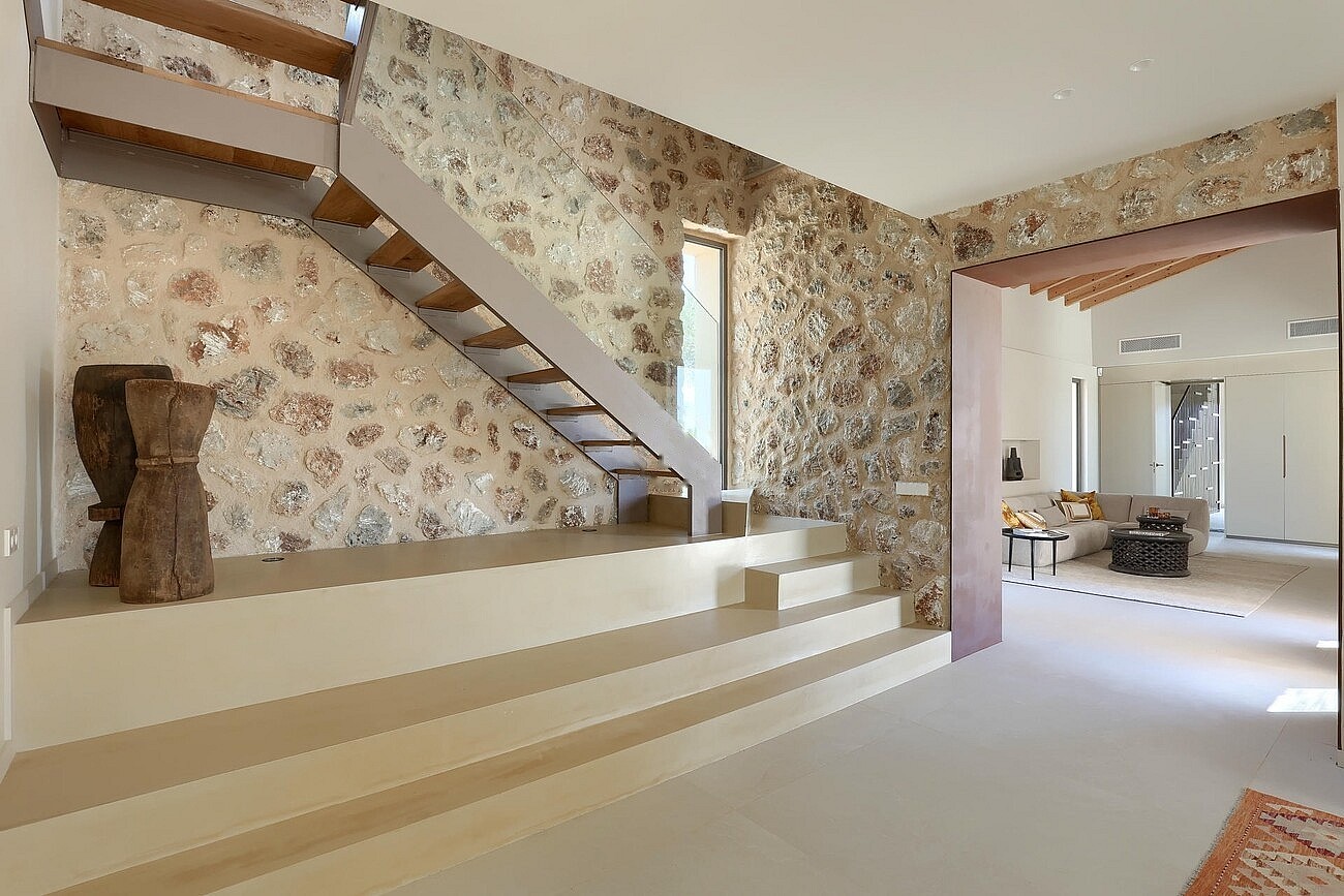 A modern staircase with wooden treads ascends against a feature wall made of exposed natural stone in a bright finca interior.