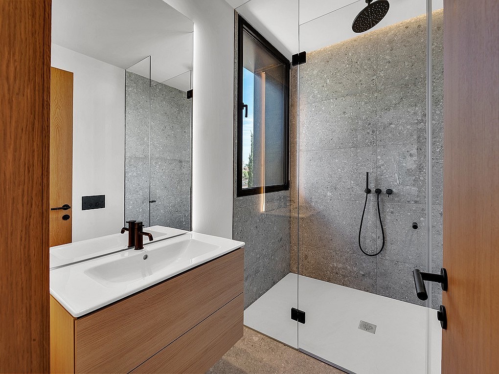 Interior view of a modern bathroom featuring a white vanity with wooden cabinetry, a large wall mirror, and a walk-in shower with grey stone tiles and black fixtures.