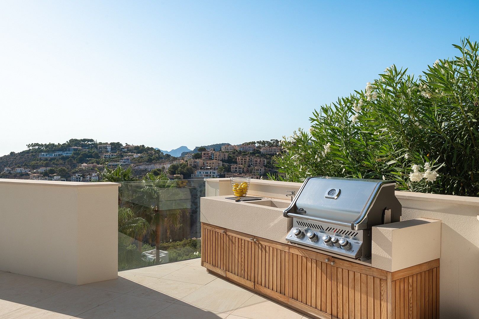 A stainless steel barbecue grill integrated into a light-toned stone and wood kitchen unit on a sunny residential terrace.