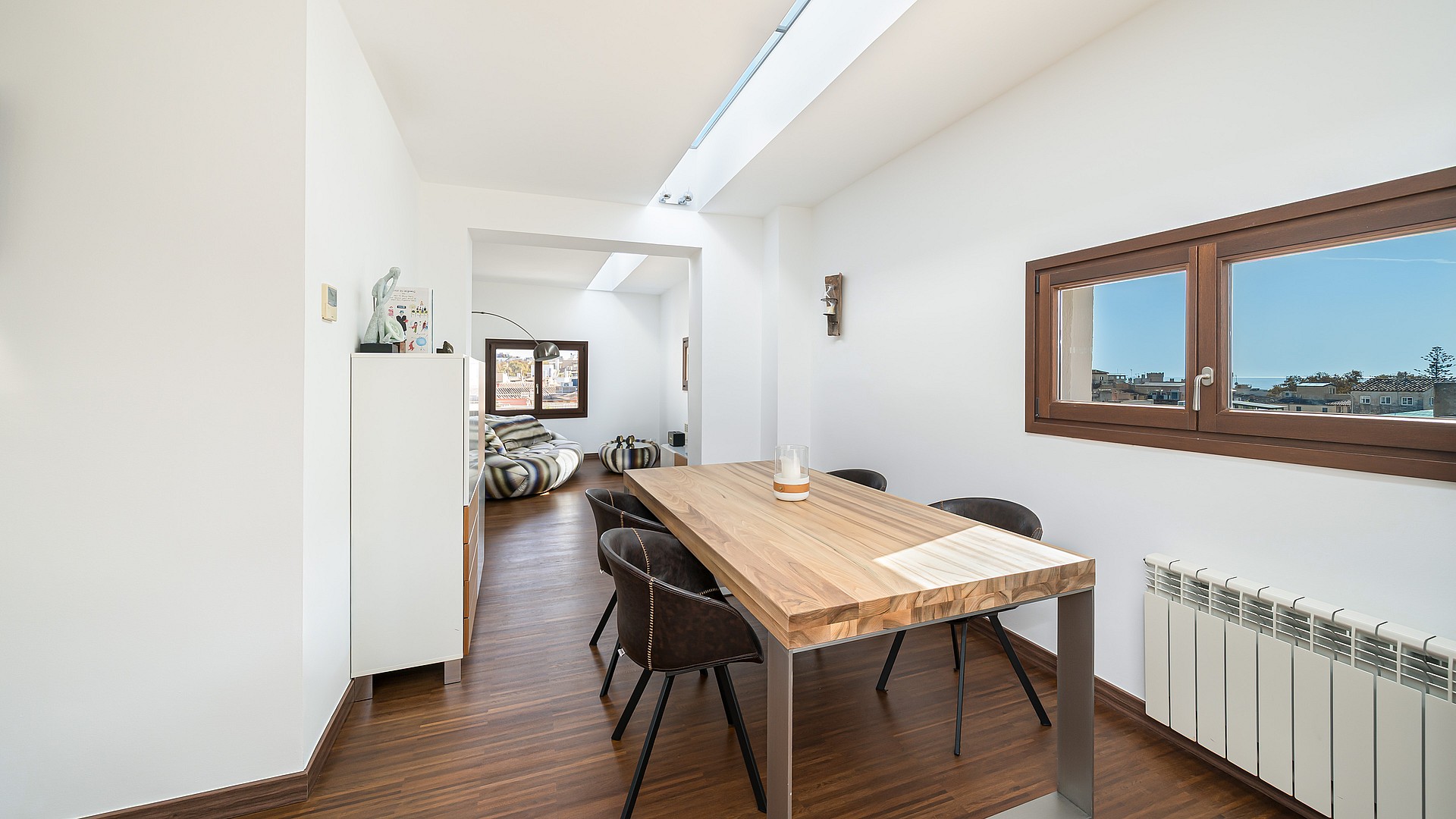 Modern dining area with oak table, dark chairs, and a long linear skylight over dark wood floors.