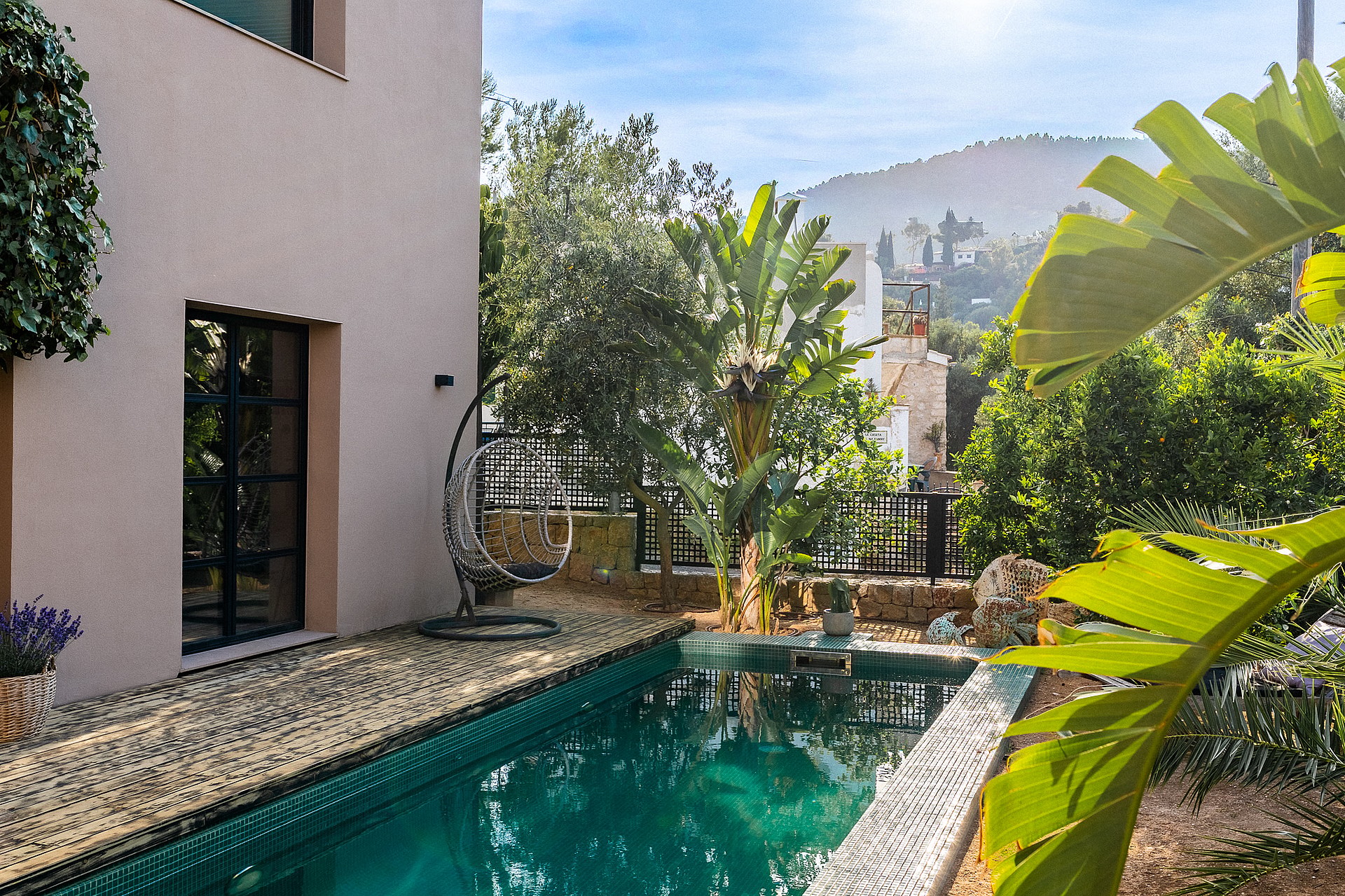 Outdoor swimming pool with turquoise water and pebble deck adjacent to a beige villa with lush green garden and mountain views.