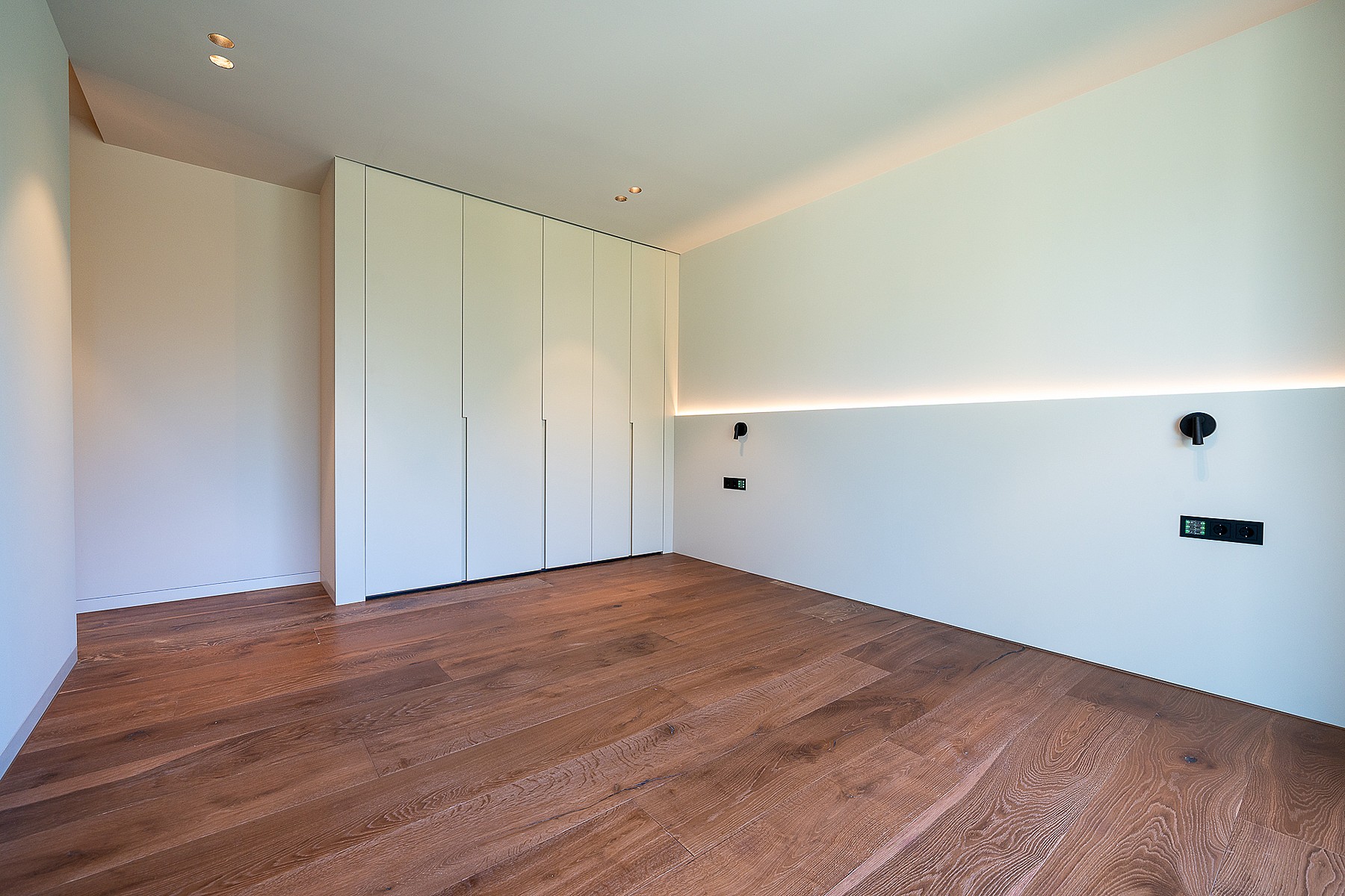 A minimalist bedroom featuring light-toned walls, oak wood flooring, and built-in floor-to-ceiling closets.