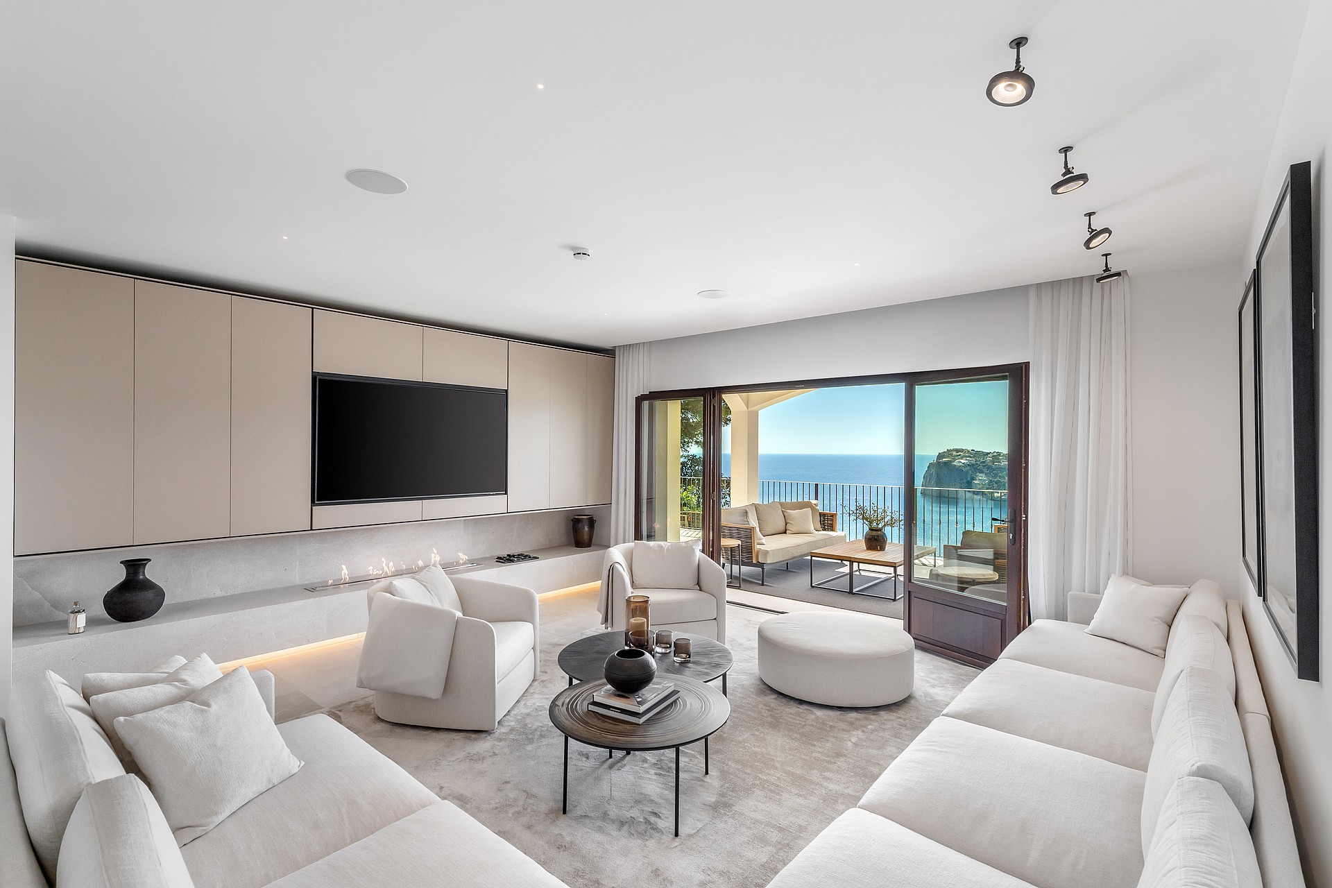 Modern white living room with soft seating and circular tables facing wide glass doors and a coastal view.