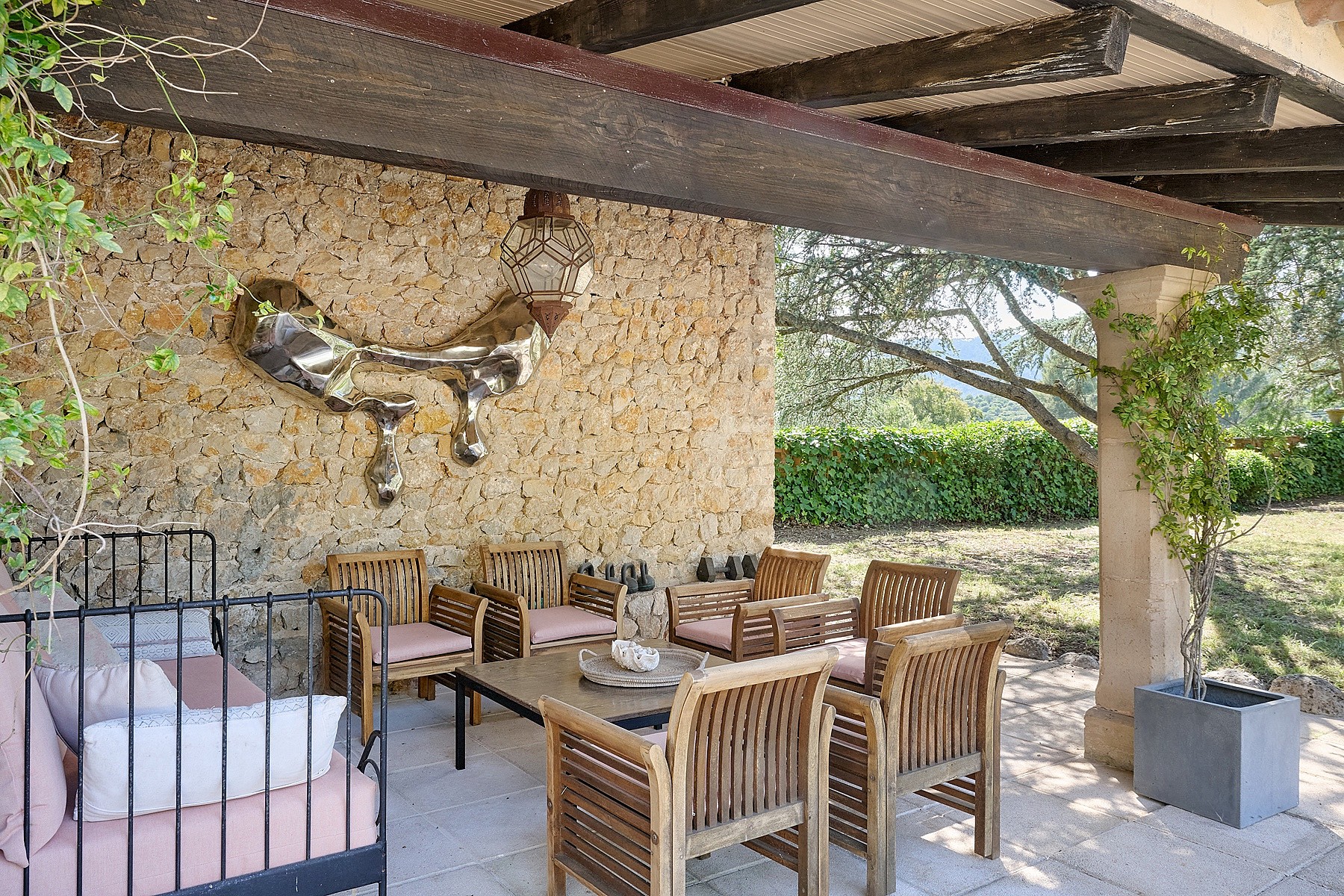 A shaded outdoor terrace with a natural stone wall, teak furniture, and a metal sculpture, overlooking greenery.