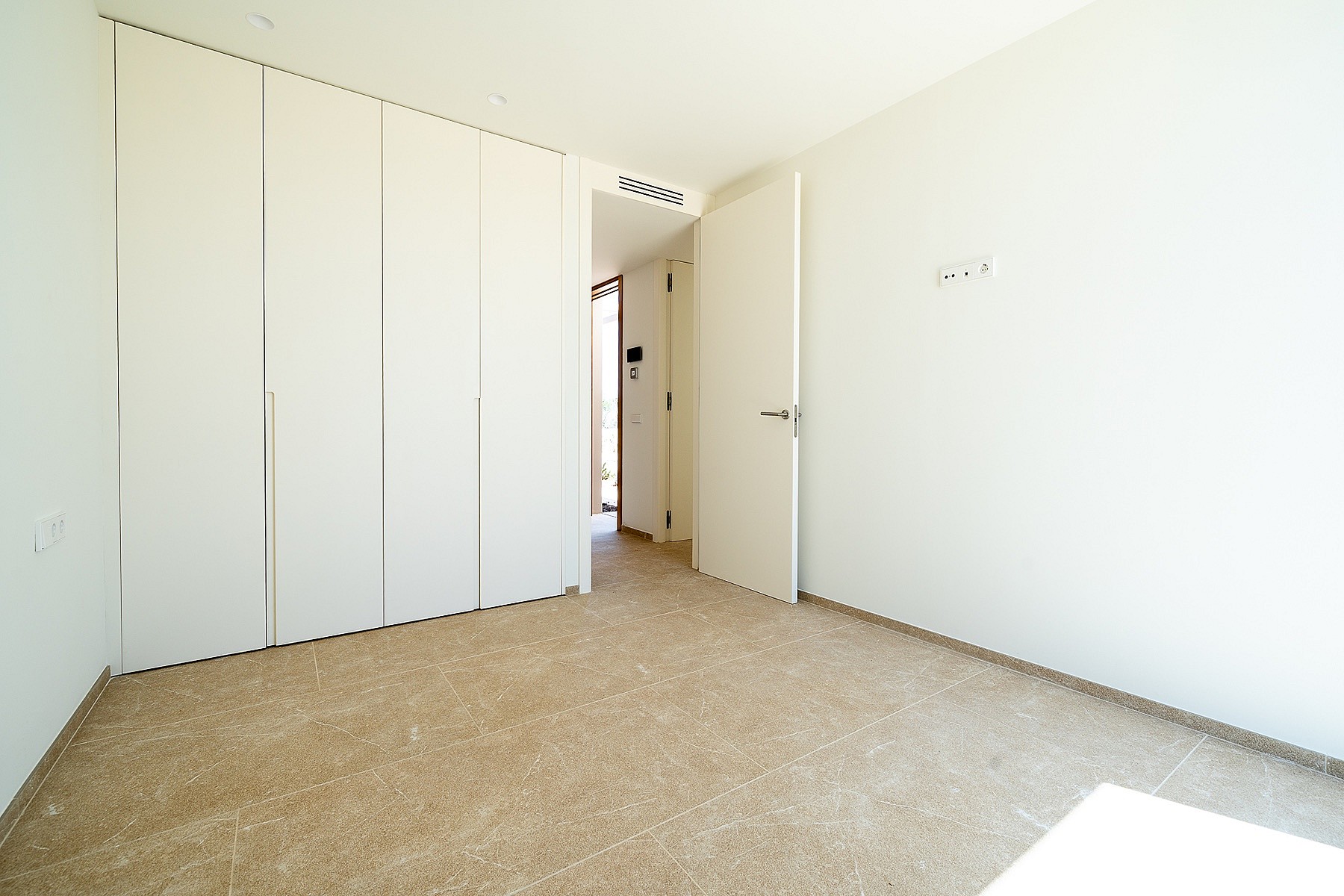 An empty bedroom featuring white built-in closets, plain walls, and beige carpet flooring with a door opening to a hallway.