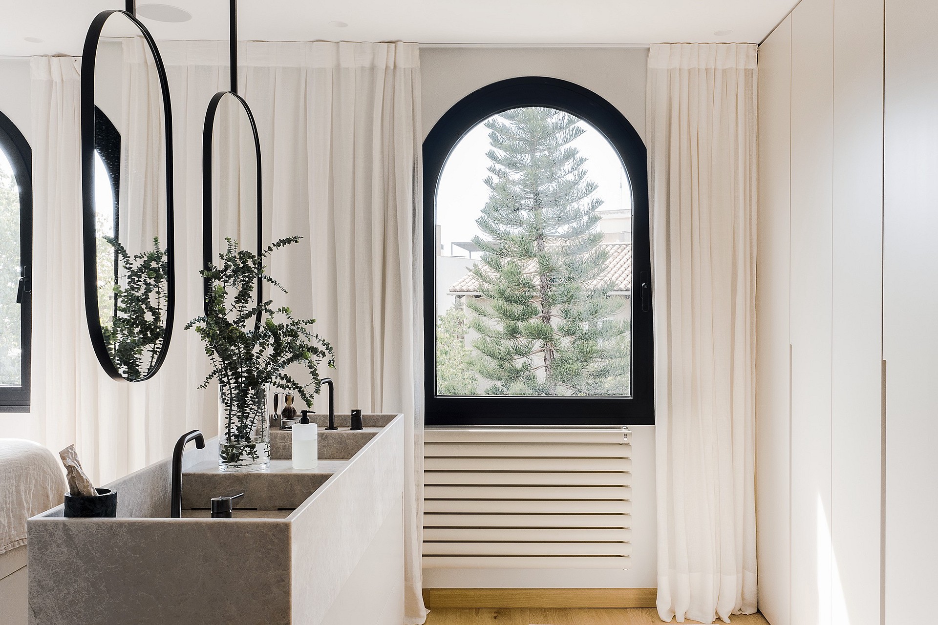 Zwei Waschbecken aus Stein mit schwarzen Armaturen, zwei ovale Hängespiegel reflektieren das Bogenfenster und Pinienausblick