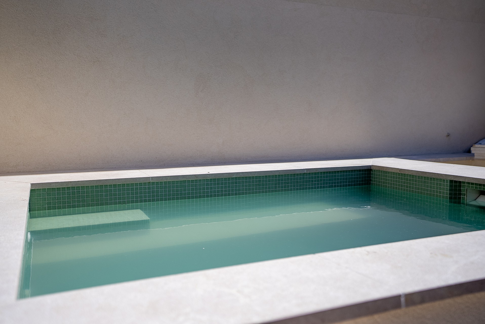 A rectangular plunge pool featuring green mosaic tiles and light-colored stone coping against a minimalist white wall.
