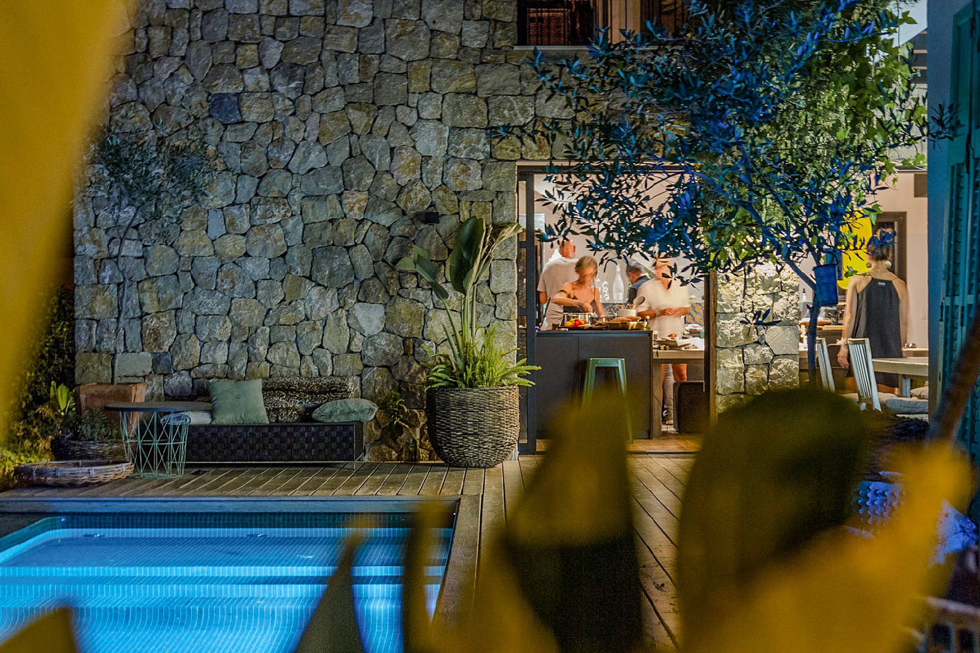 A stone-walled outdoor terrace with a swimming pool and a view into a lit interior kitchen, framed by foreground foliage.