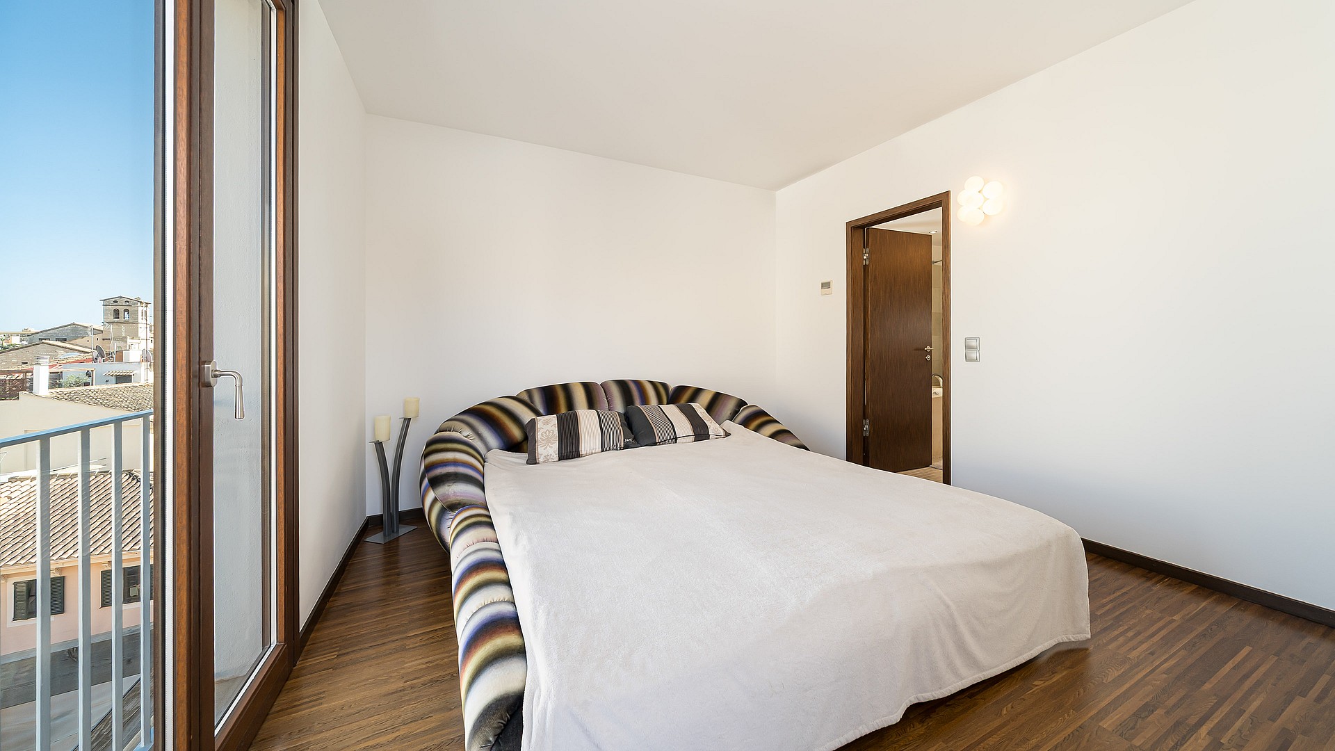 Modern bedroom featuring dark hardwood floors and a French balcony overlooking the historic rooftops of Palma.