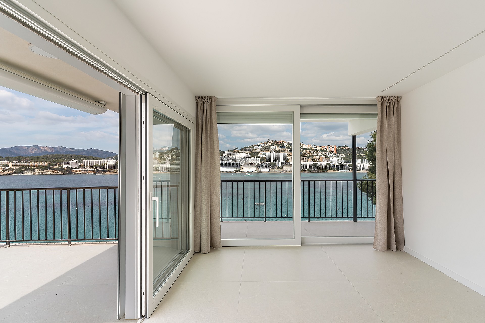 An interior view of large sliding glass doors opening onto a balcony with a black railing, overlooking a bay and hills.