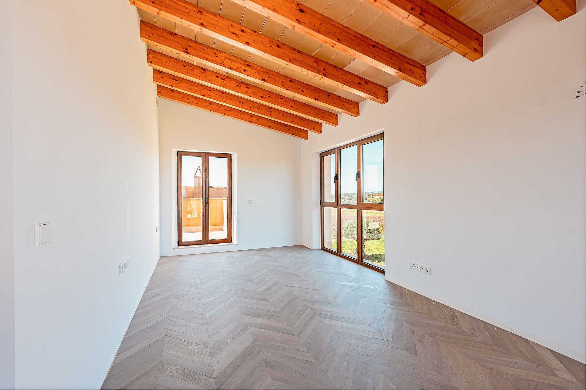 An empty, bright bedroom with white walls, herringbone parquet flooring, and exposed wooden ceiling beams, featuring large windows.