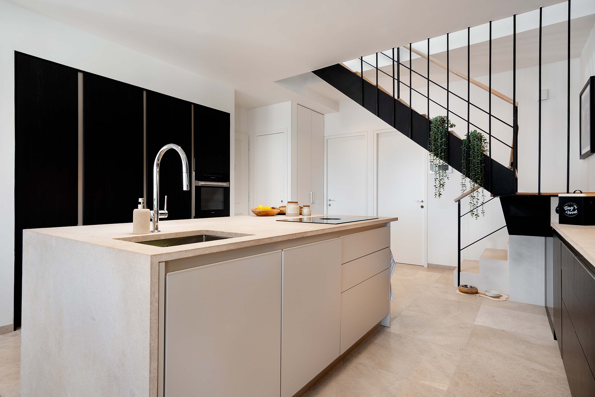 Modern kitchen with a light stone island, matte black cabinetry, and a floating staircase with thin metal railings.