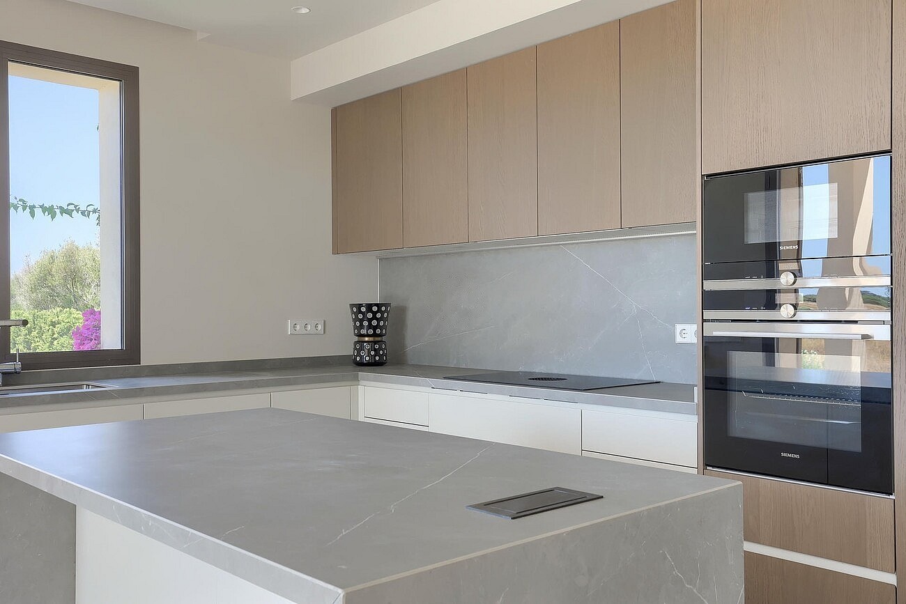A modern kitchen with a grey stone-topped island, light wood cabinetry, and a double wall oven next to a window.