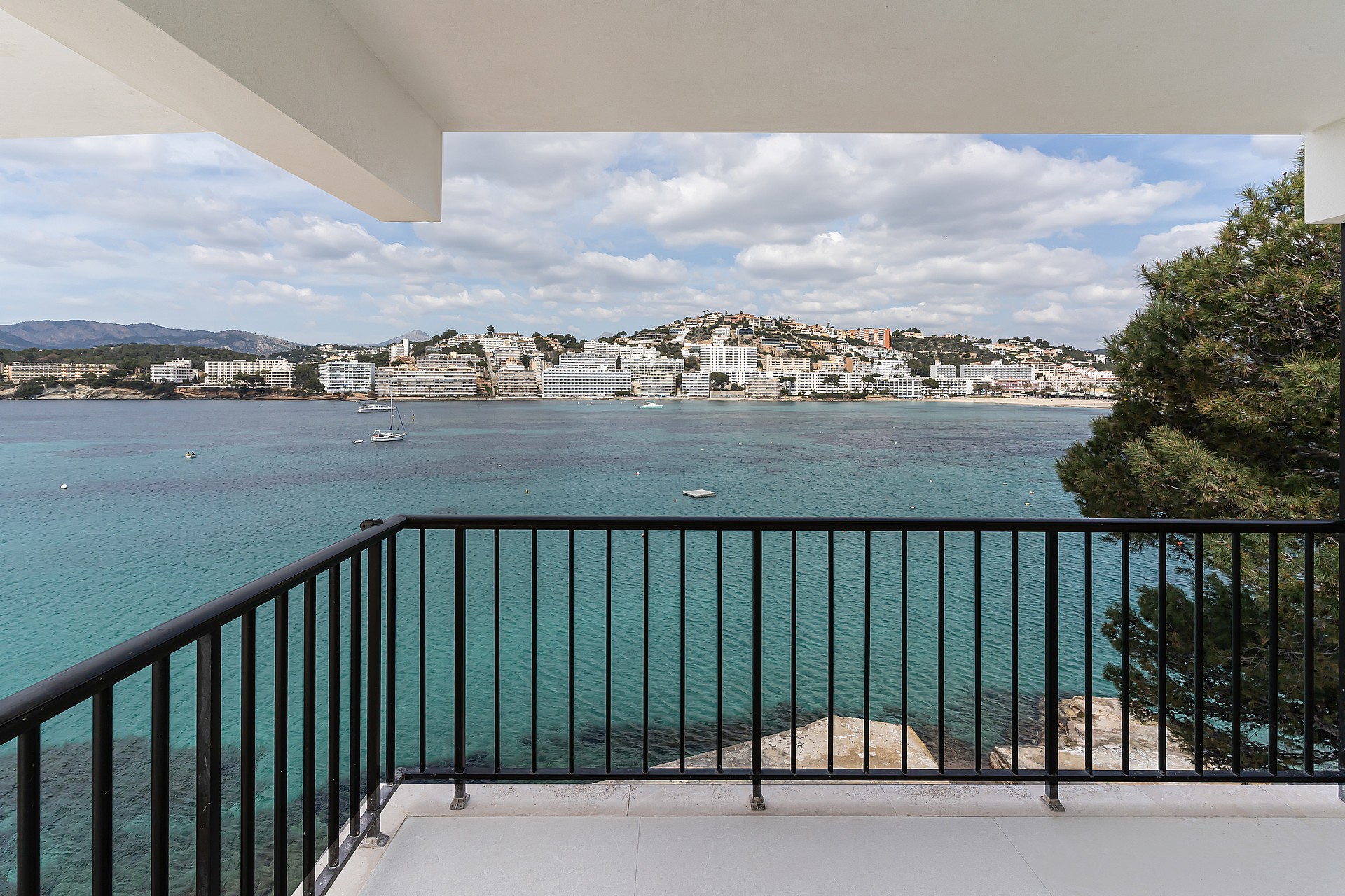 A balcony with black metal railing overlooking a turquoise bay and white coastal town under a soft sky.