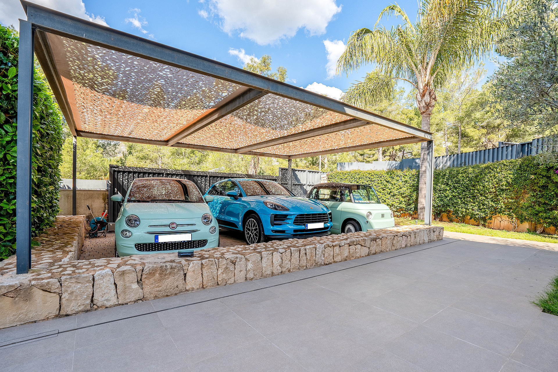 Covered carport with steel frame and patterned shade canopy beside a natural-stone wall.
