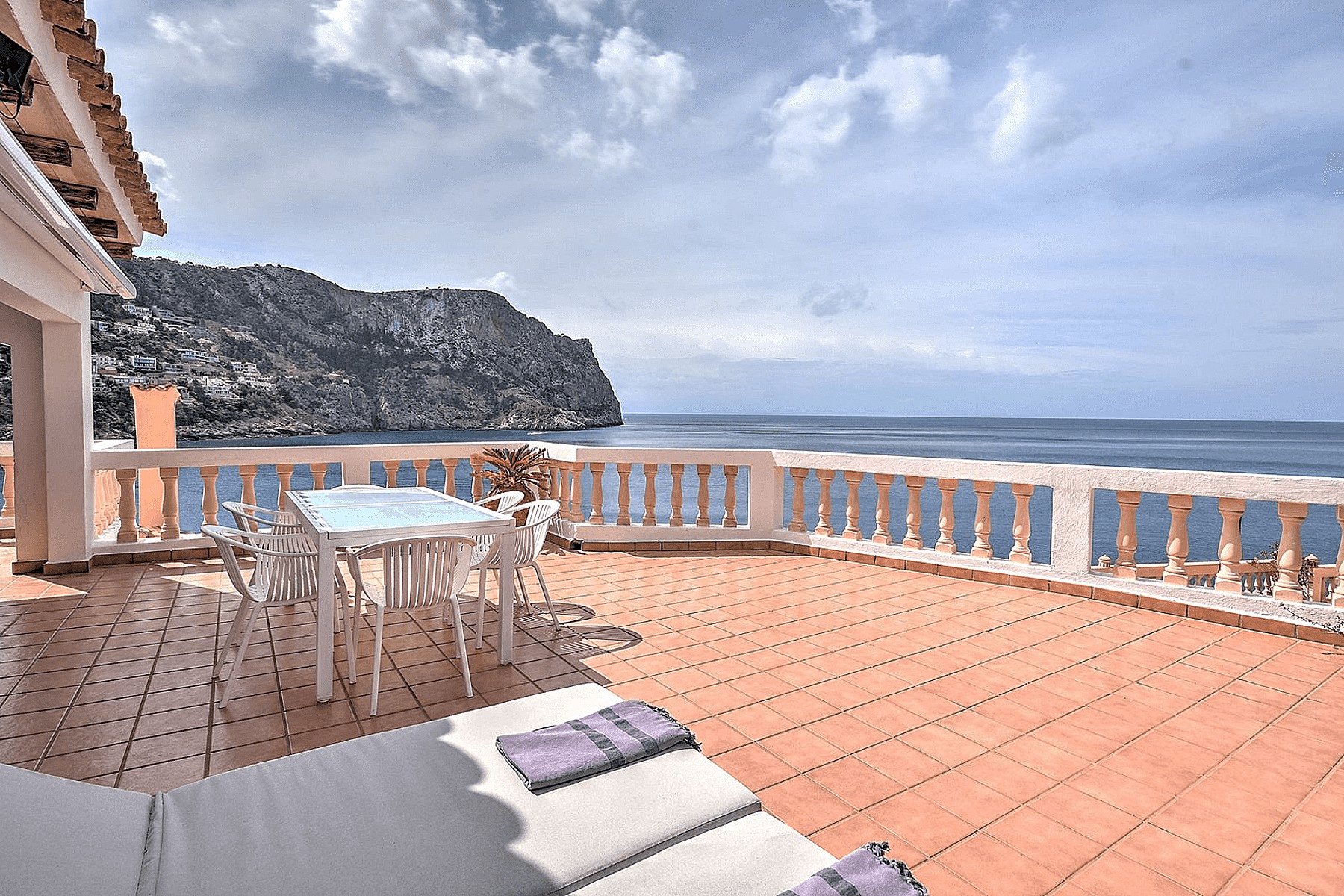 A wide outdoor terrace in Cala Llamp with terracotta floor tiles, a stone balustrade, and white outdoor dining furniture overlooking the sea.