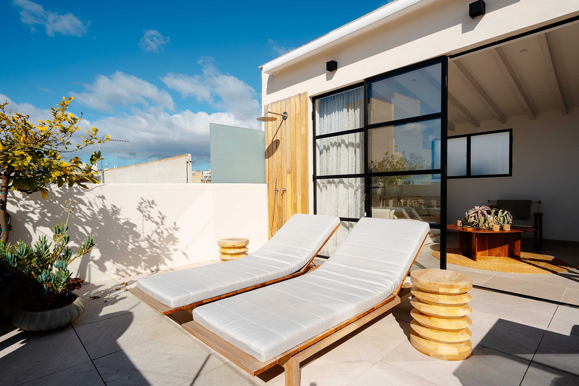 A private terrace in Santa Catalina featuring two wooden lounge chairs, a wall-mounted shower, and a black-framed glass door.