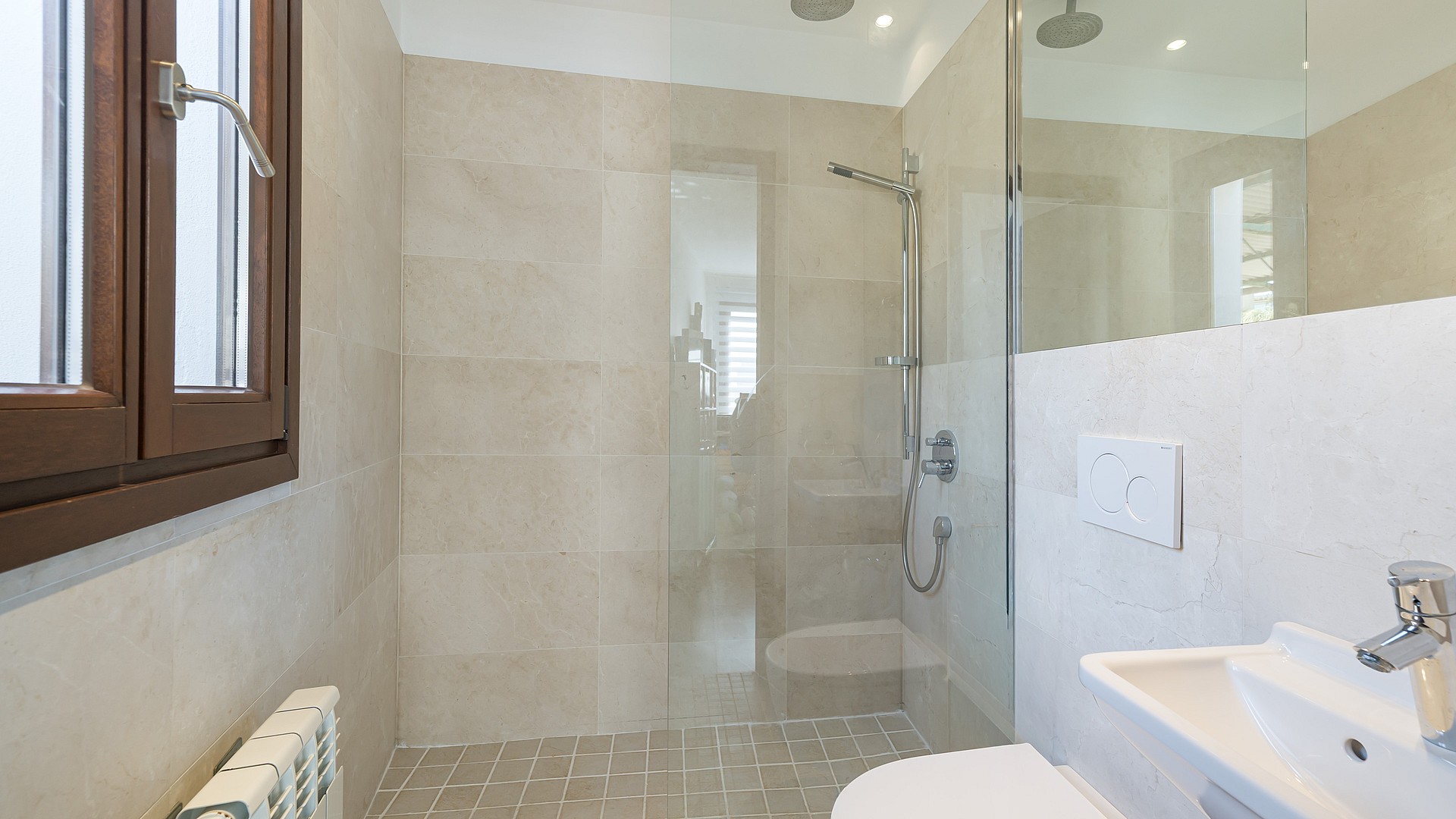 Contemporary bathroom with beige tiling, glass walk-in shower, rainfall head, and a wooden Mallorcan-style window.