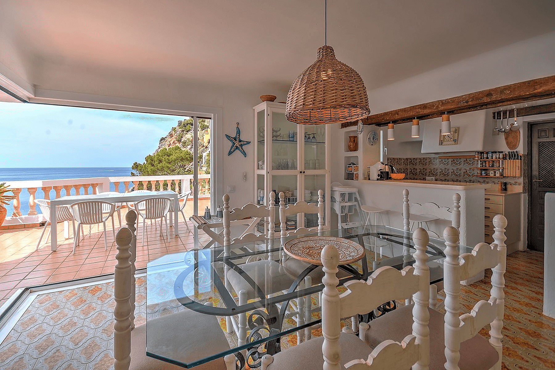 Interior dining room with a glass-top table, white wooden chairs, and a rattan pendant light, opening to a terrace with sea views.