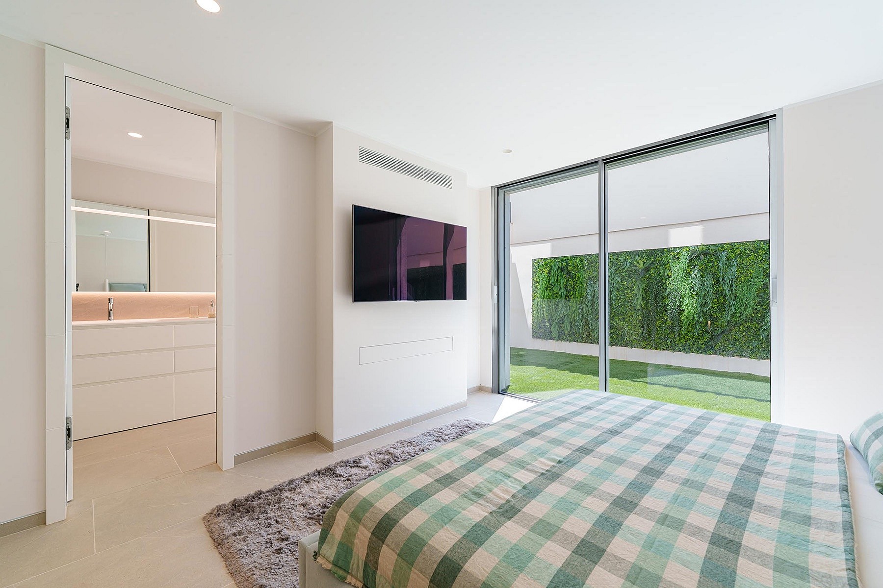 Contemporary bedroom with checkered bedspread, wall-mounted television, and sliding glass doors opening to a green wall.