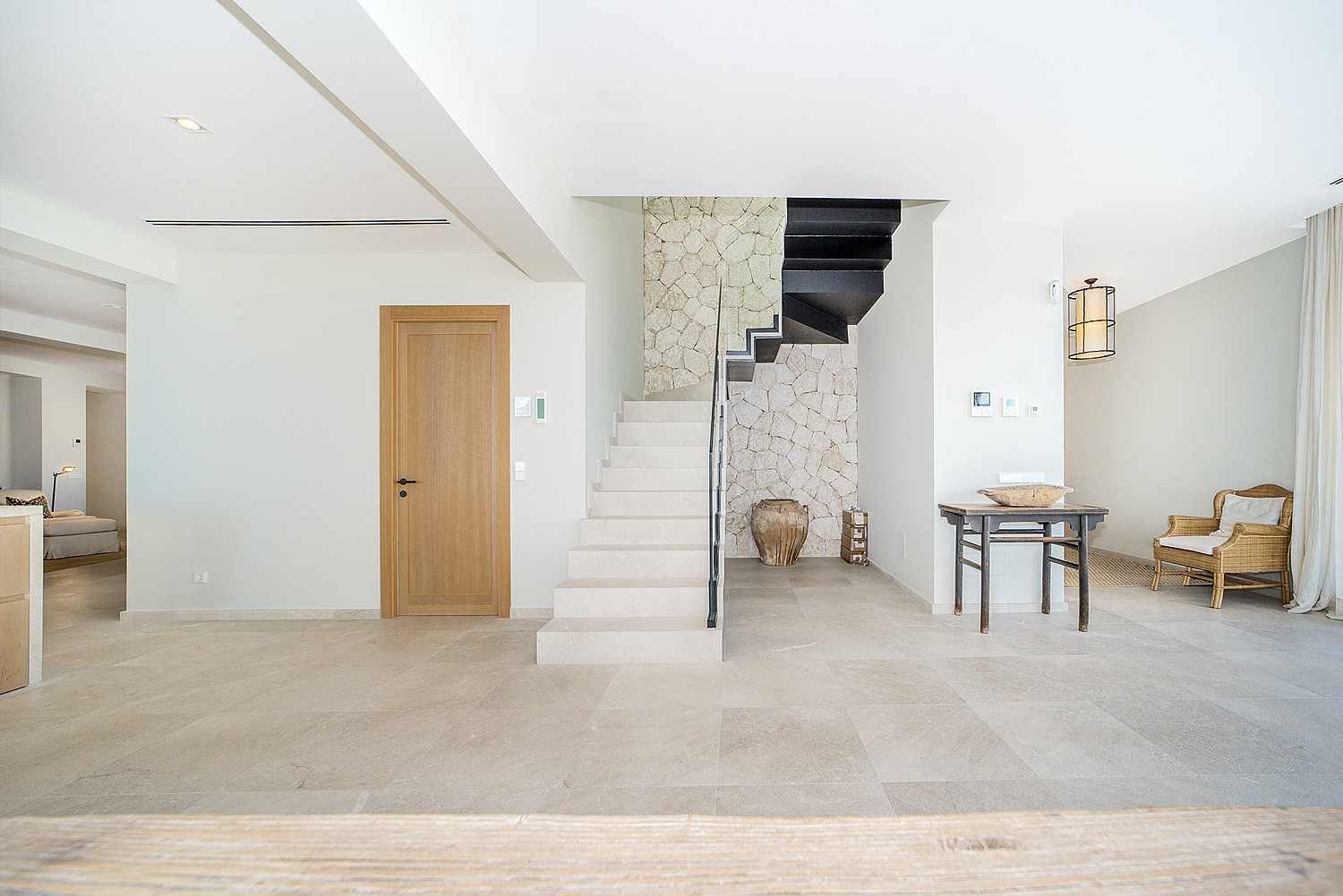 A wide entrance hall featuring a minimalist white stone staircase, a light oak wood door, and a textured stone feature wall.