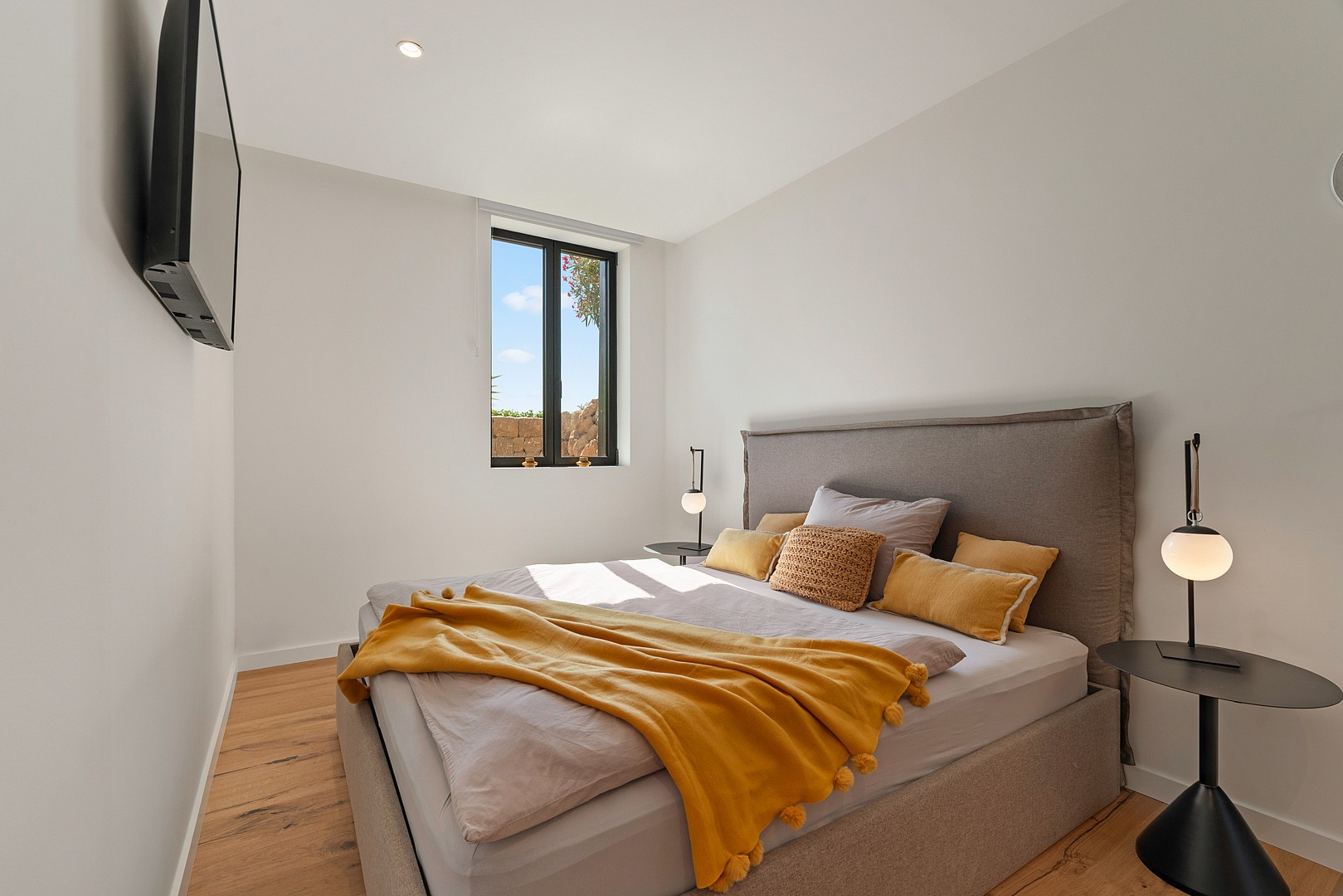 A modern bedroom with an upholstered grey bed, mustard yellow throw blanket, wooden flooring, and a single vertical window.