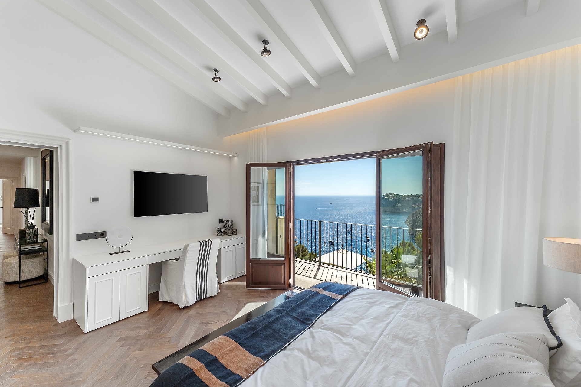 Master bedroom featuring white beamed ceilings, herringbone wood floors, and a balcony opening to Mediterranean sea views.