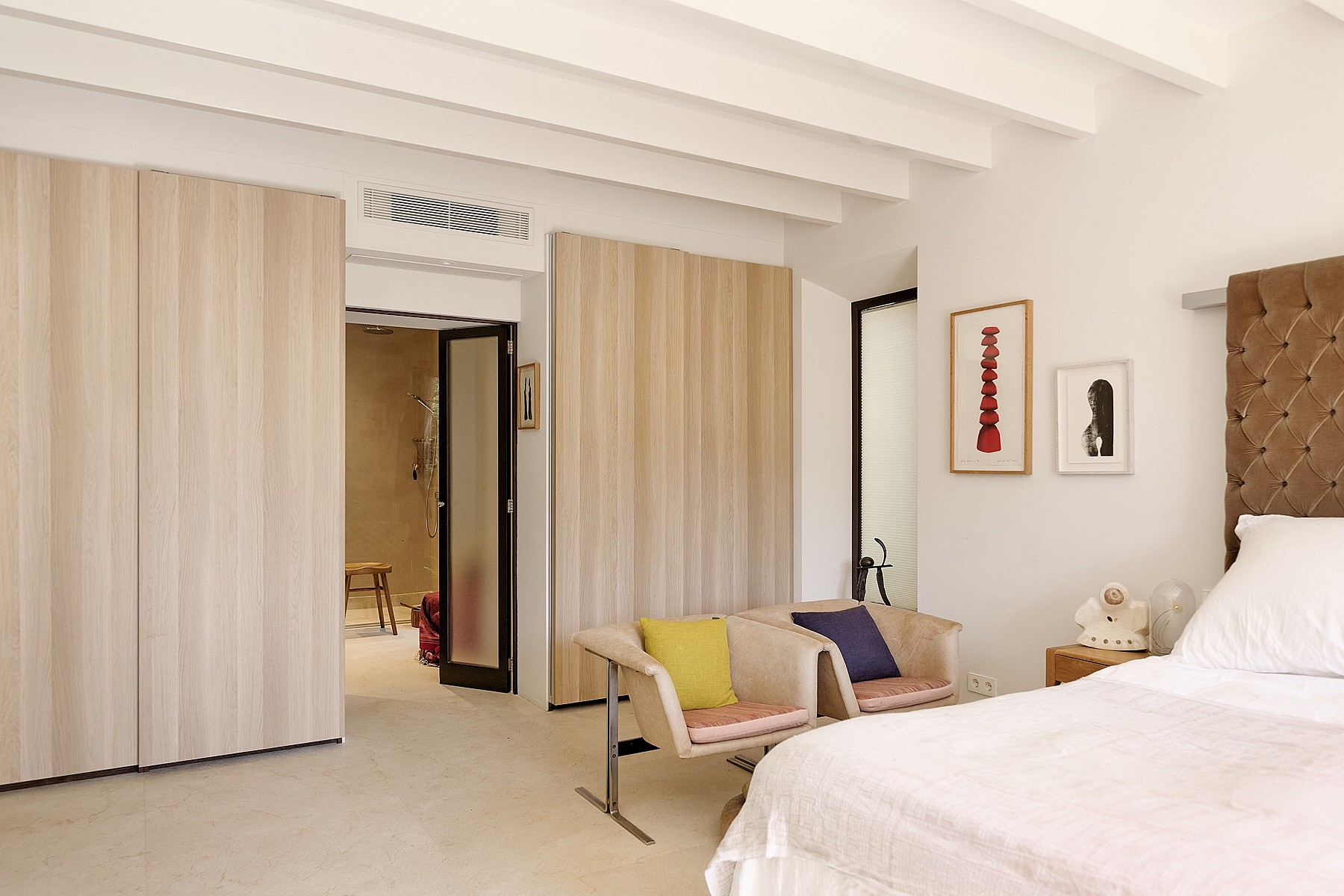 A bright bedroom with white beamed ceiling, light wood wardrobe doors, and a partial view into an en-suite bathroom.