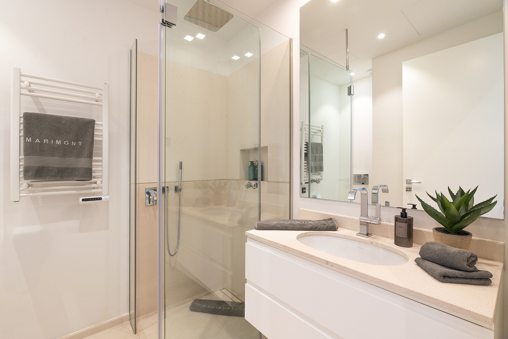Modern bathroom featuring a white vanity with cream stone top, glass-enclosed walk-in shower, and heated towel rail.
