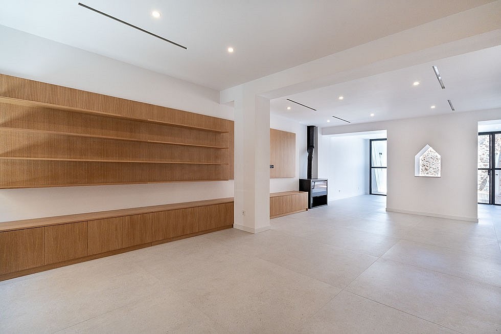 A bright, minimalist open-plan living room with light-colored stone floors, white walls, and long, wall-mounted oak shelving units.