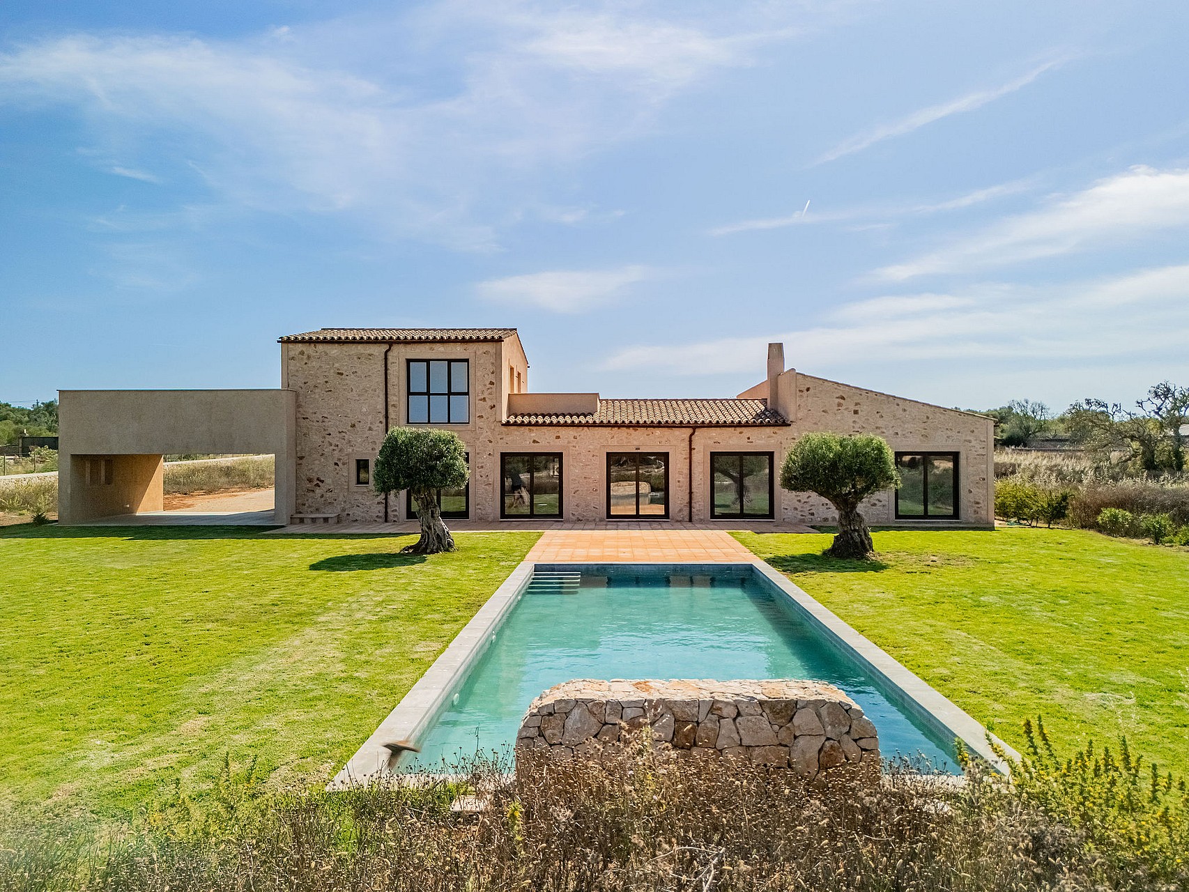 Exterior view of a stone finca in Santanyí featuring a rectangular swimming pool, manicured lawn, and large glass doors.