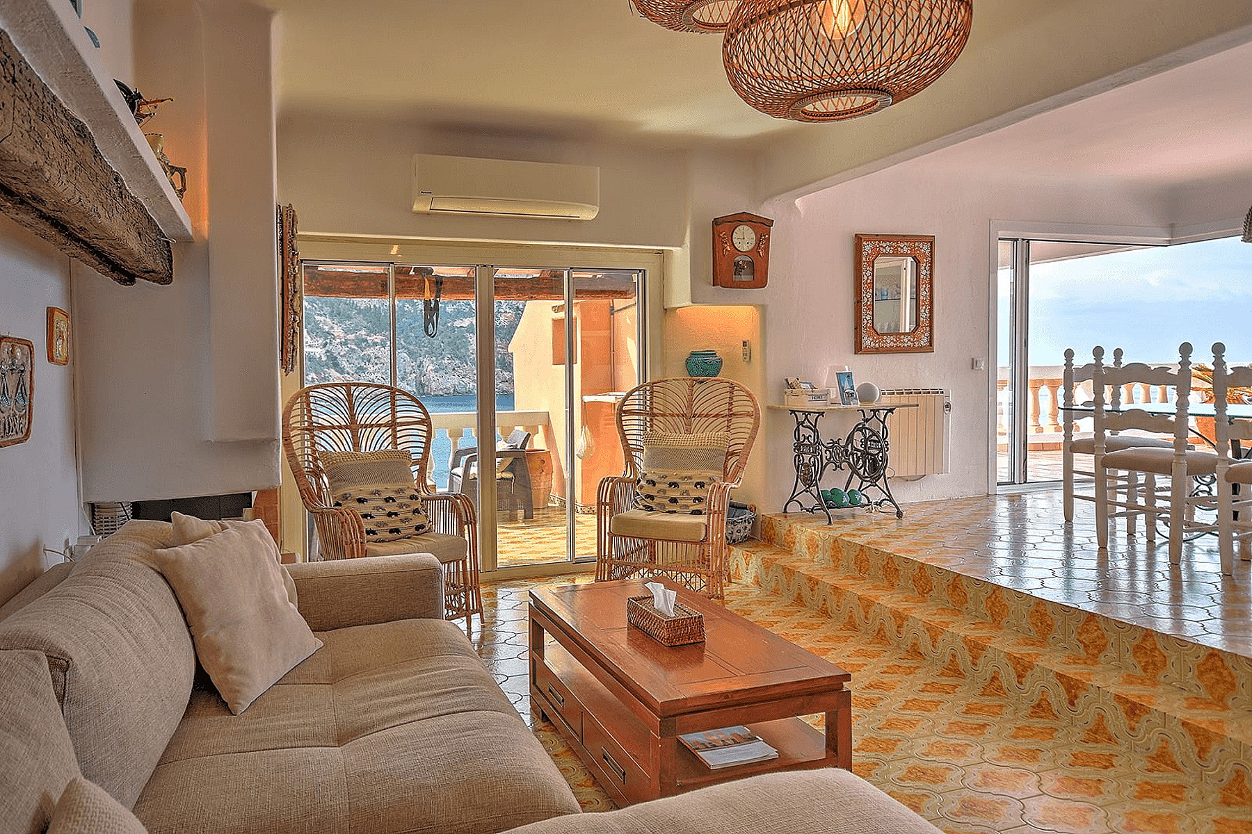 Sunlit living area with tiled floors, rattan armchairs, and a view of Cala Llamp through glass sliding doors.
