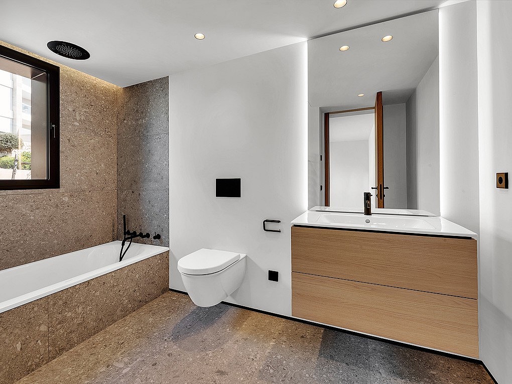 Bathroom interior featuring a wall-mounted toilet, light wood vanity, and brown stone tile bathtub surround with matte black fixtures.