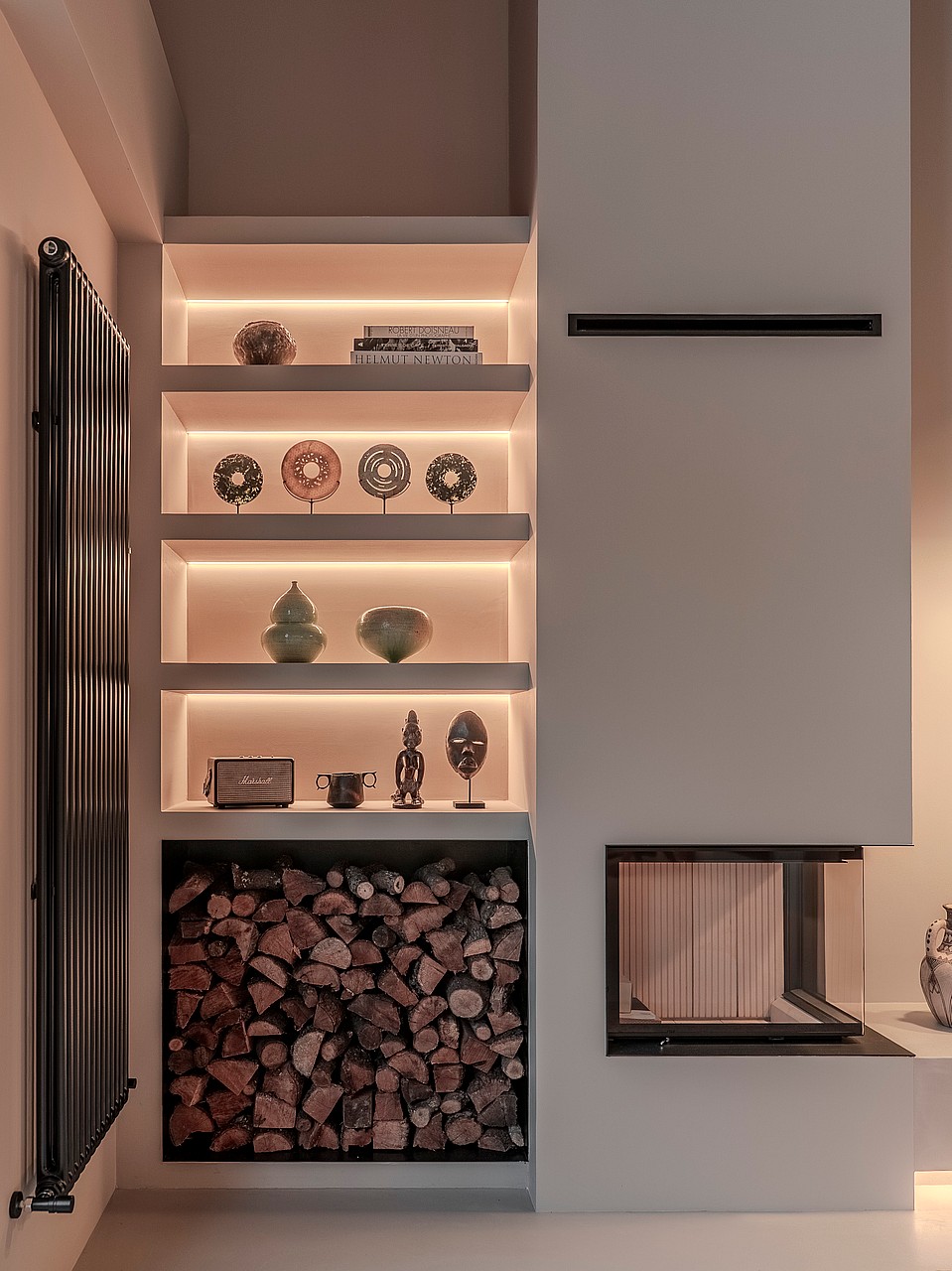 A modern white fireplace with a glass corner unit, recessed backlit shelves displaying artifacts, and a log storage niche.