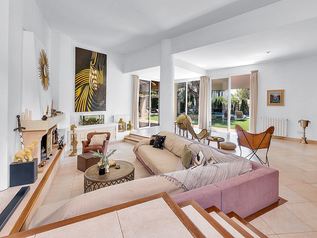 A bright, open-plan living room with a large corner sofa, a fireplace, and glass doors leading to an outdoor garden area.