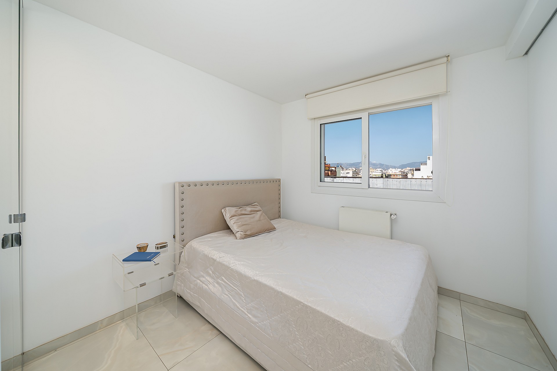 A minimalist bedroom featuring a neutral upholstered bed, white walls, and a window framing an urban Old Town vista.