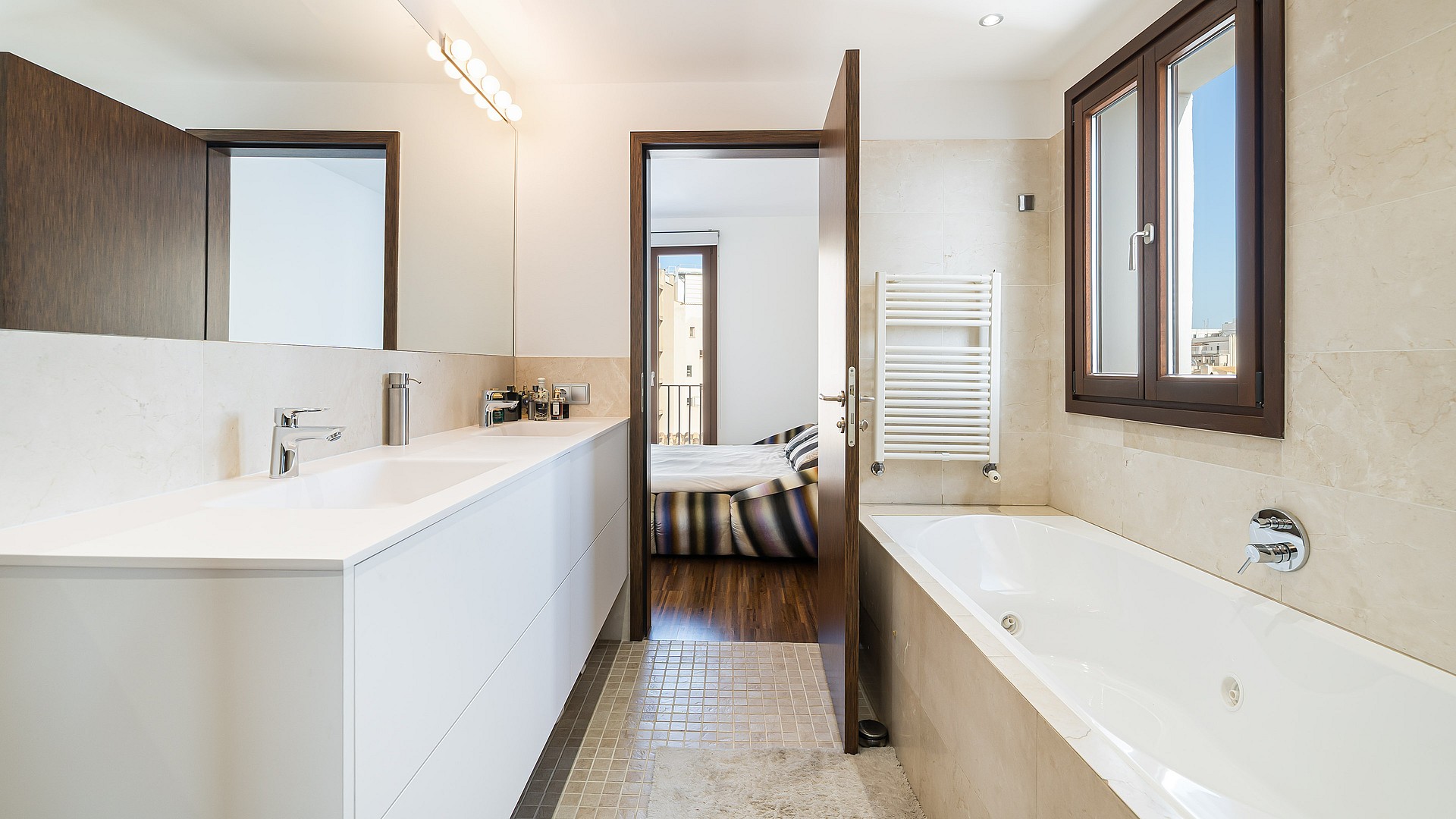 Bright bathroom featuring a white double vanity, integrated bathtub, natural stone wall tiling, and wooden door access.
