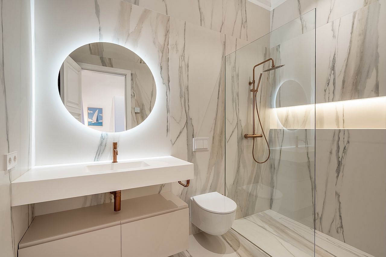 A modern bathroom featuring white marble wall tiles, a floating vanity, a backlit circular mirror, and a glass-enclosed shower.