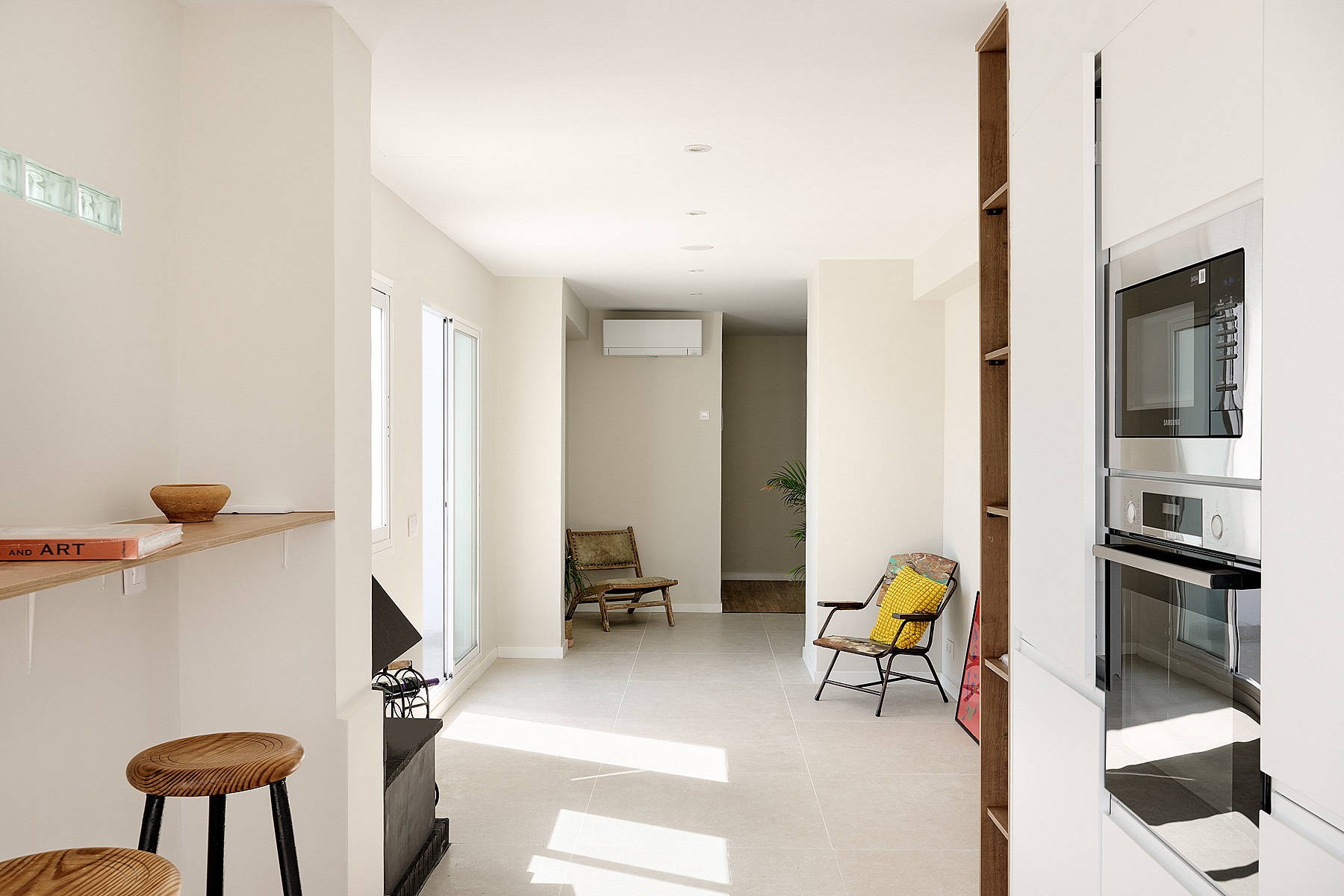 Interior view from a kitchen looking towards a living area with white walls, tiled floors, a stool, and a yellow chair.