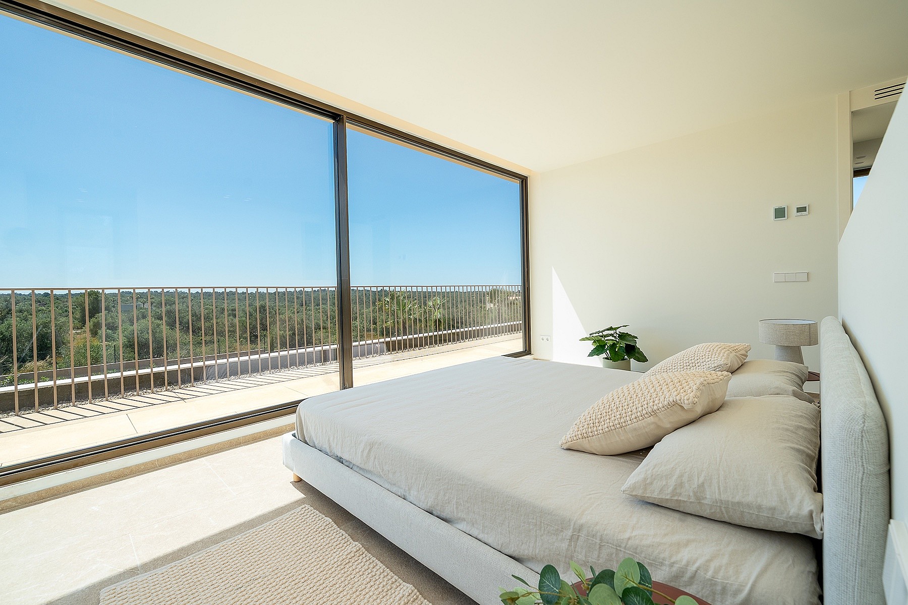 A minimalist bedroom featuring a platform bed with beige bedding, overlooking a sunlit balcony with mountain views.