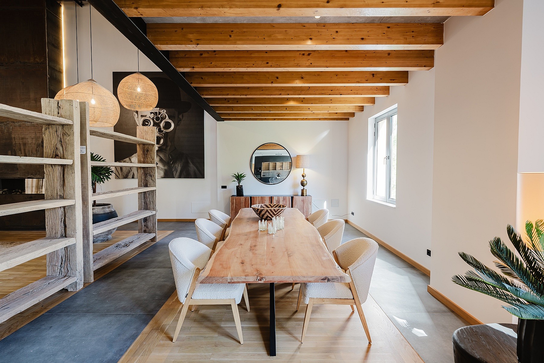 Dining area featuring a long live-edge wooden table, white upholstered chairs, and exposed wooden ceiling beams in a spacious room.