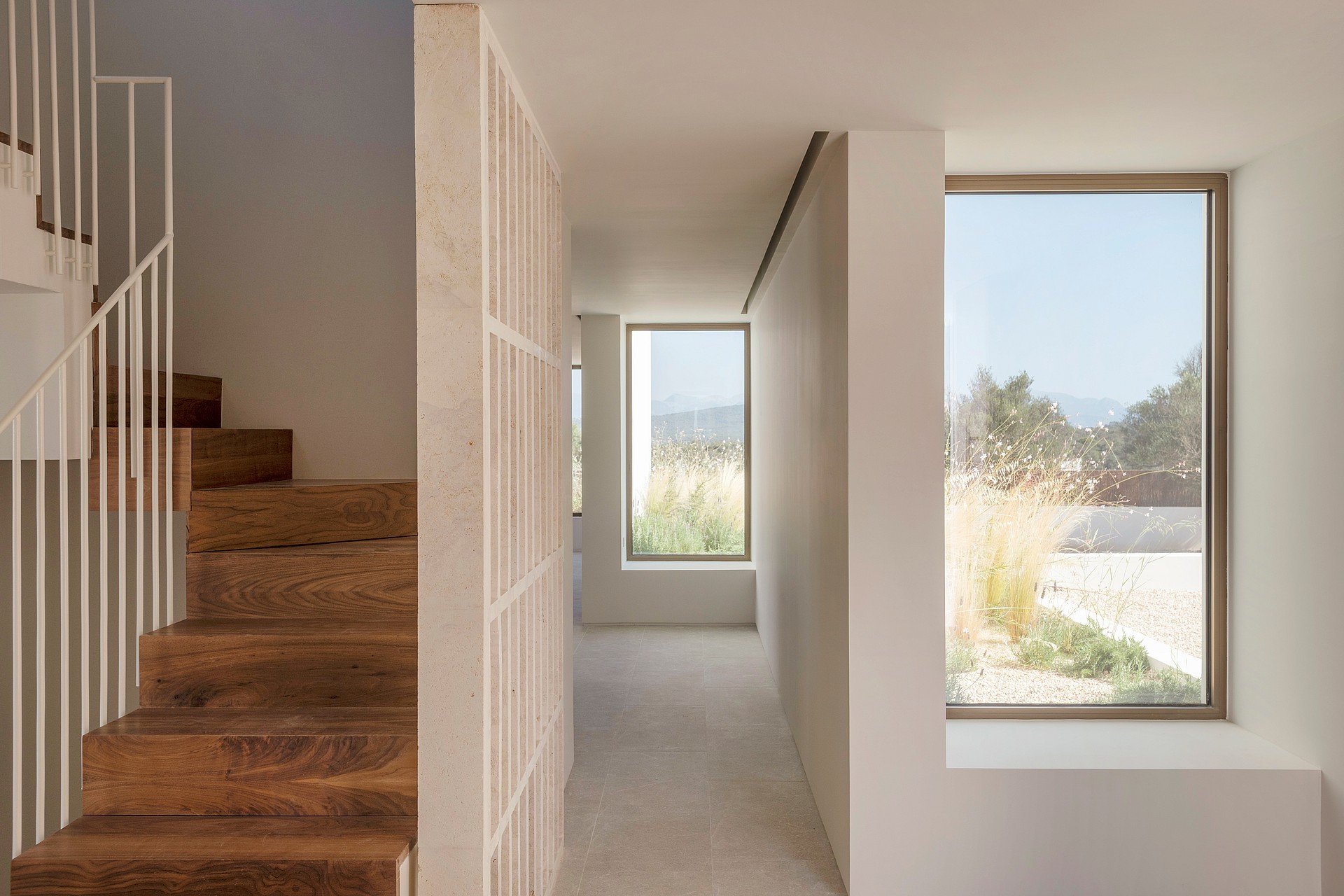 A bright minimalist hallway featuring a vertical timber screen, wooden stairs, and large windows overlooking a field.