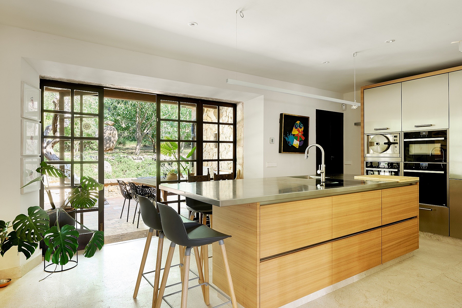 Modern kitchen with light wood cabinetry, stone-topped island, and black metal framed glass doors opening to a garden patio.